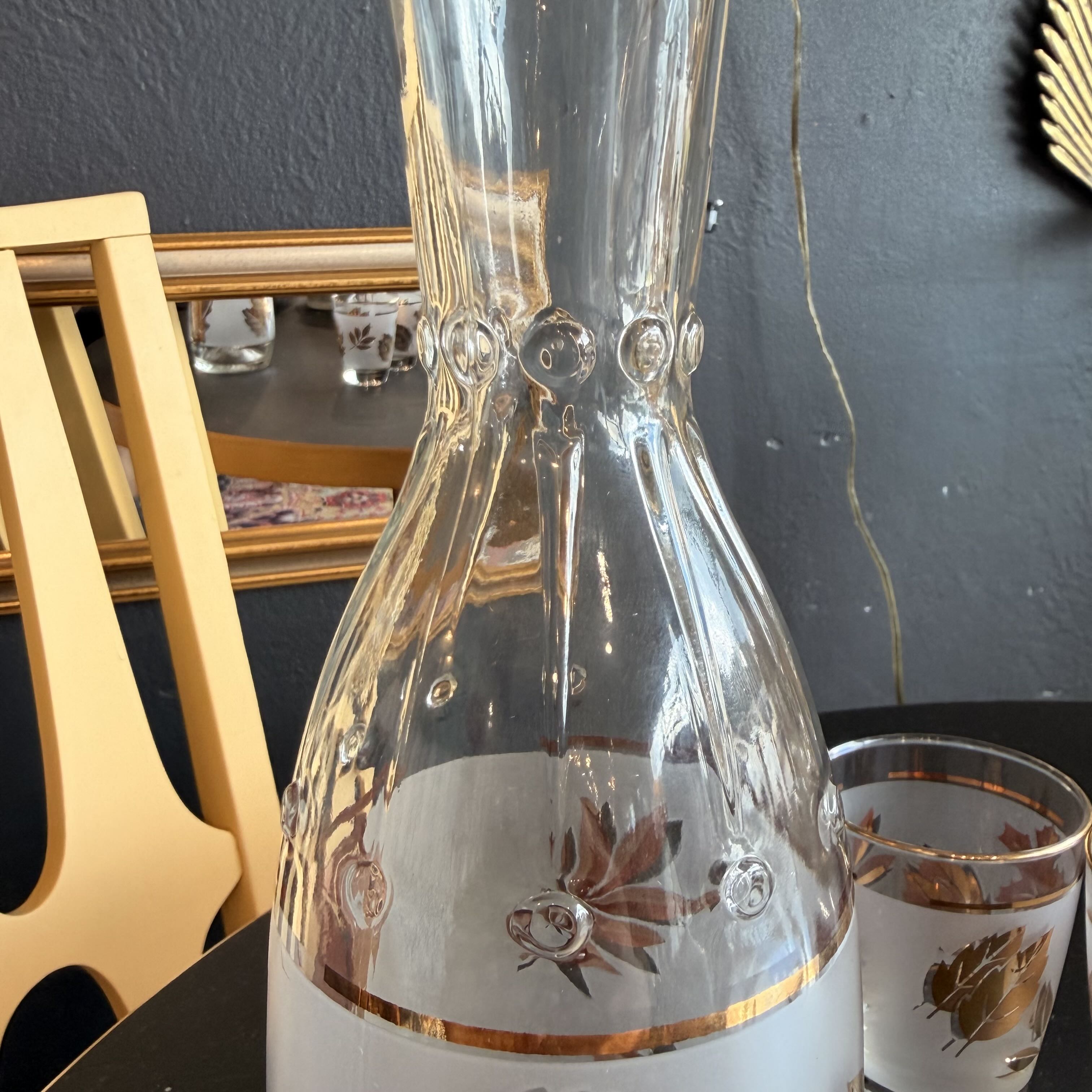 A clear glass carafe with a white lower section and gold-toned decorative leaf motifs, shown alongside matching drinking glasses on a dark table against a dark wall.