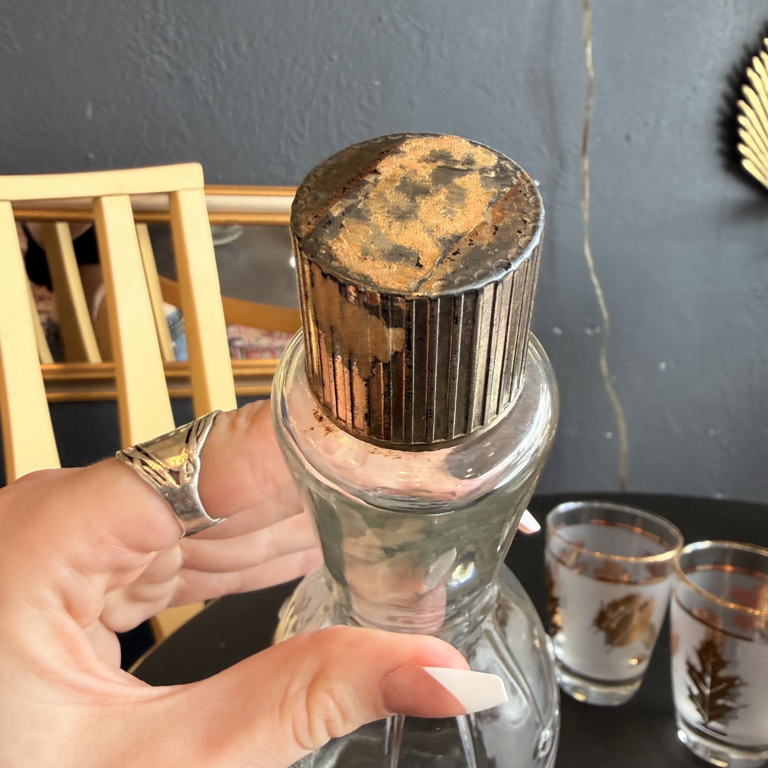 A hand holds a clear glass bottle with a ribbed metallic cap that has a heavily distressed, rusted brown finish. Two matching etched glass tumblers sit on a dark table in the background against a dark gray wall.