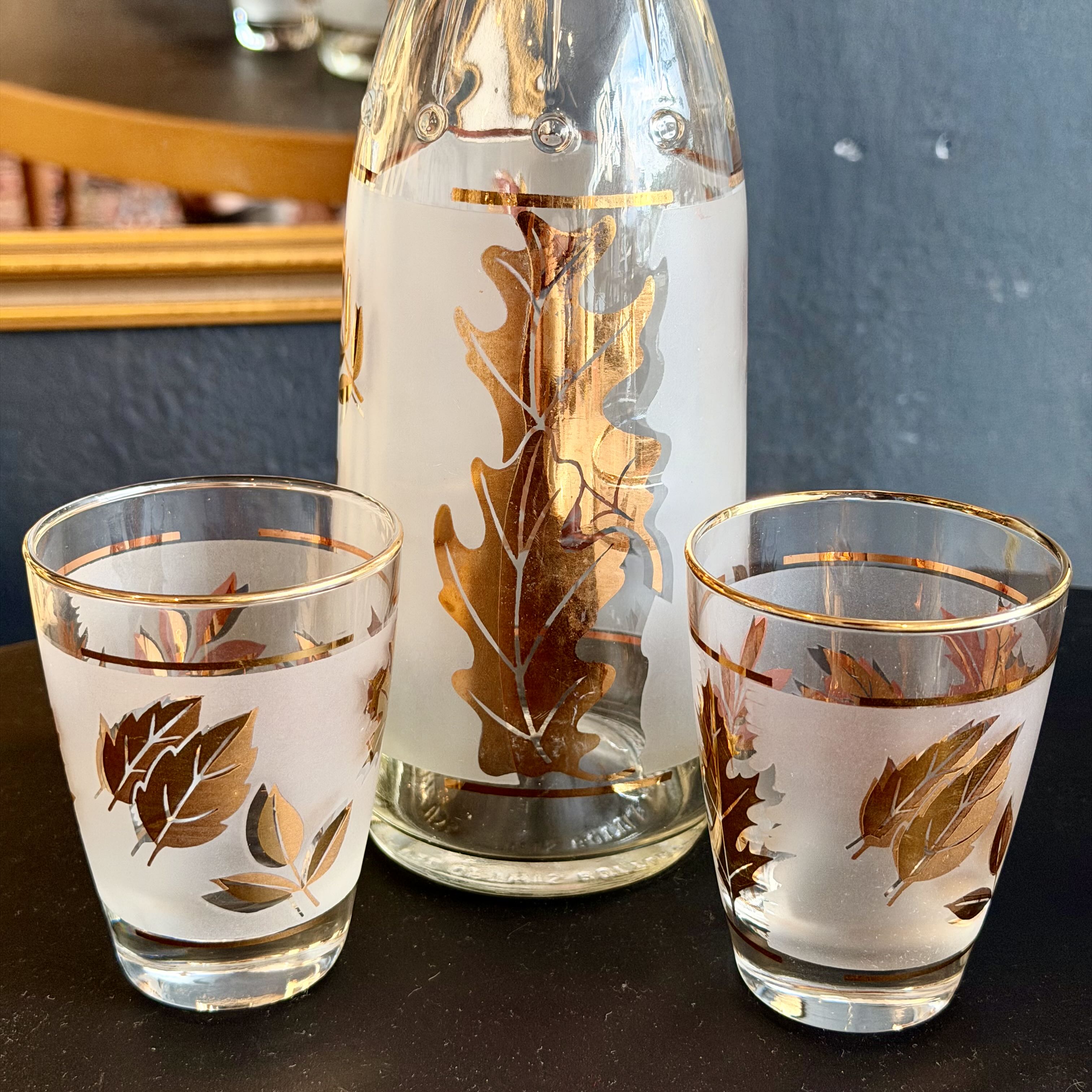 A frosted glass sake bottle and two matching shot glasses decorated with metallic gold and copper-toned oak leaf patterns, set on a dark tabletop.