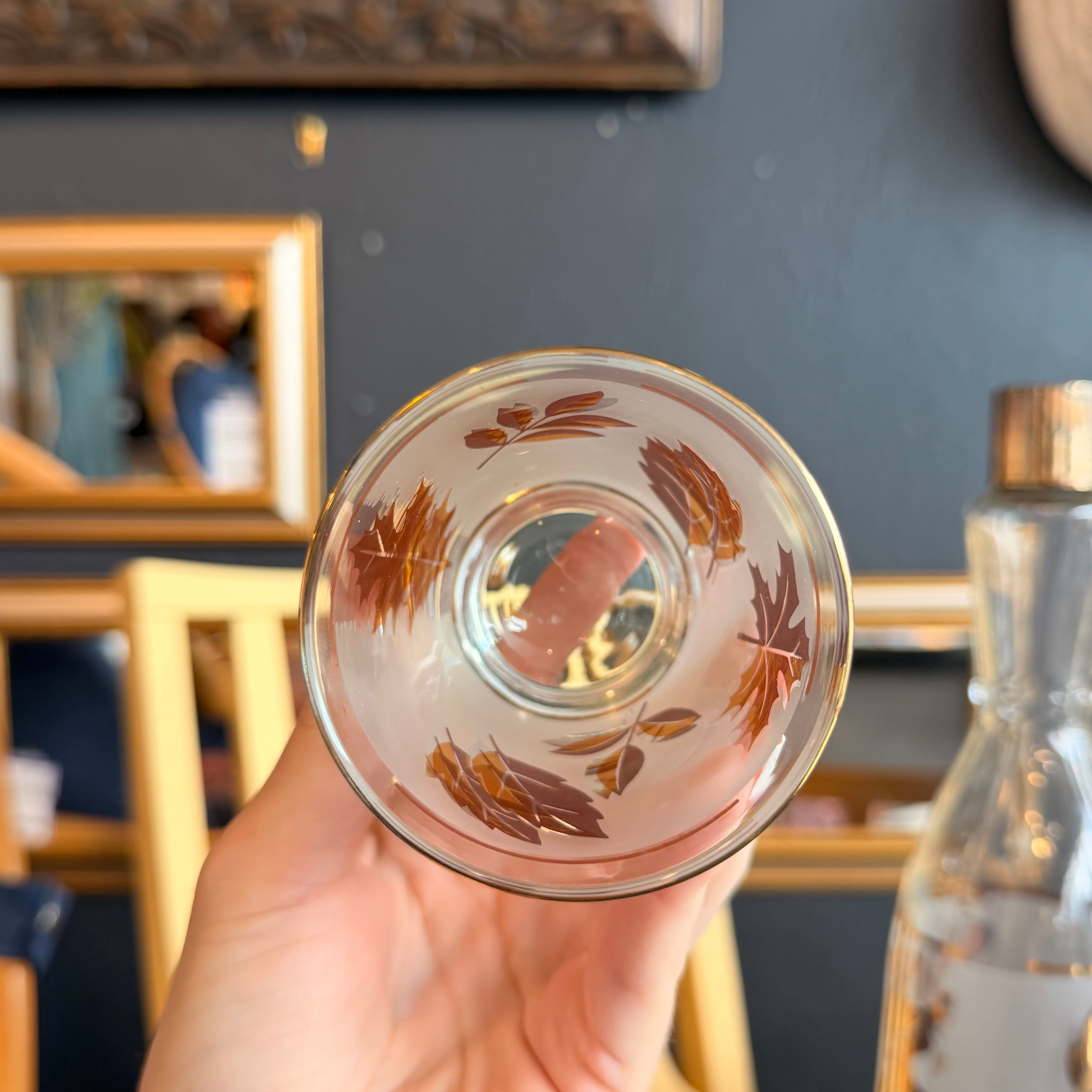 A hand with white manicured nails holds a frosted glass tumbler decorated with metallic gold leaf patterns and gold trim, set against a blurred home interior background.