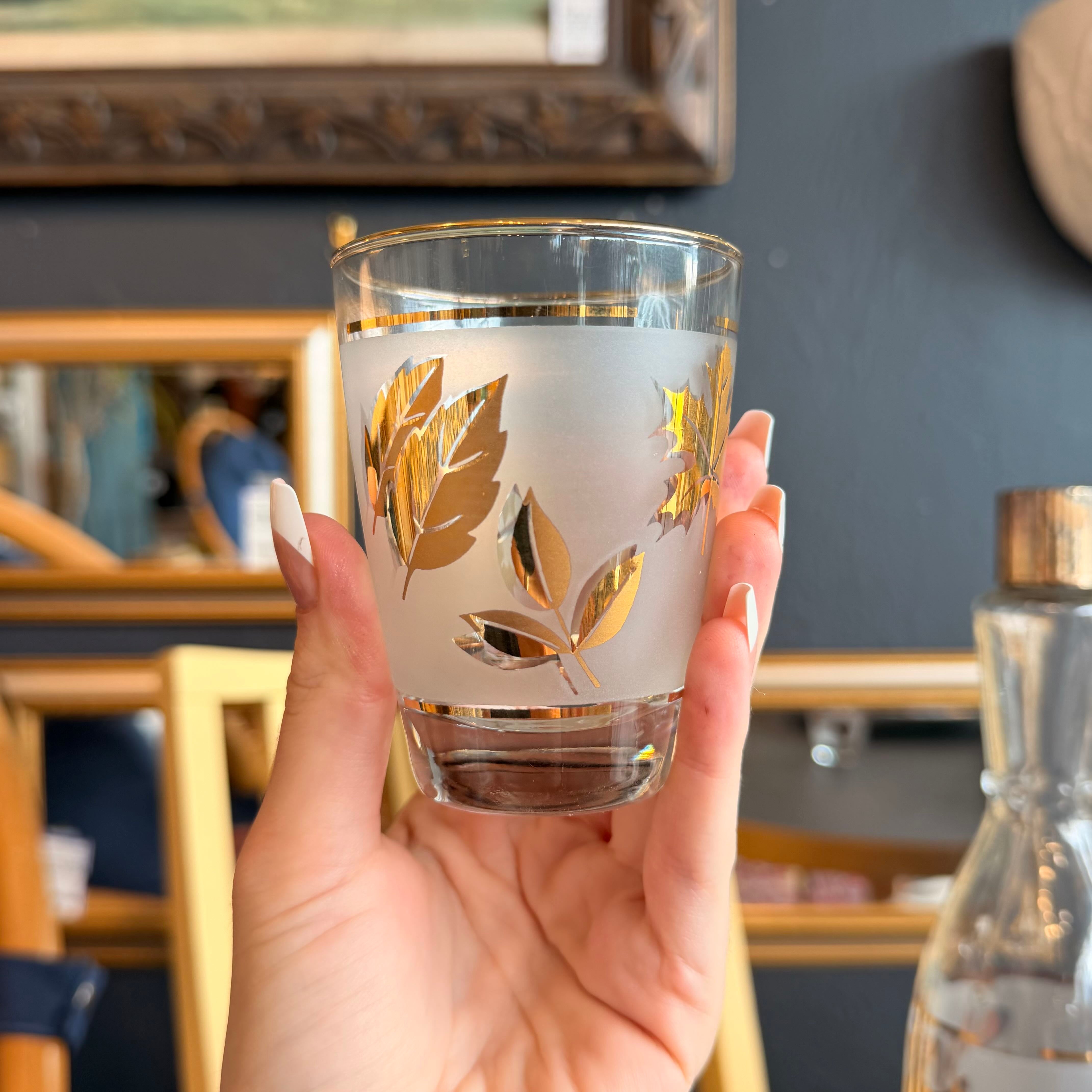 A hand with white manicured nails holds a frosted glass tumbler decorated with metallic gold leaf patterns and gold trim, set against a blurred home interior background.