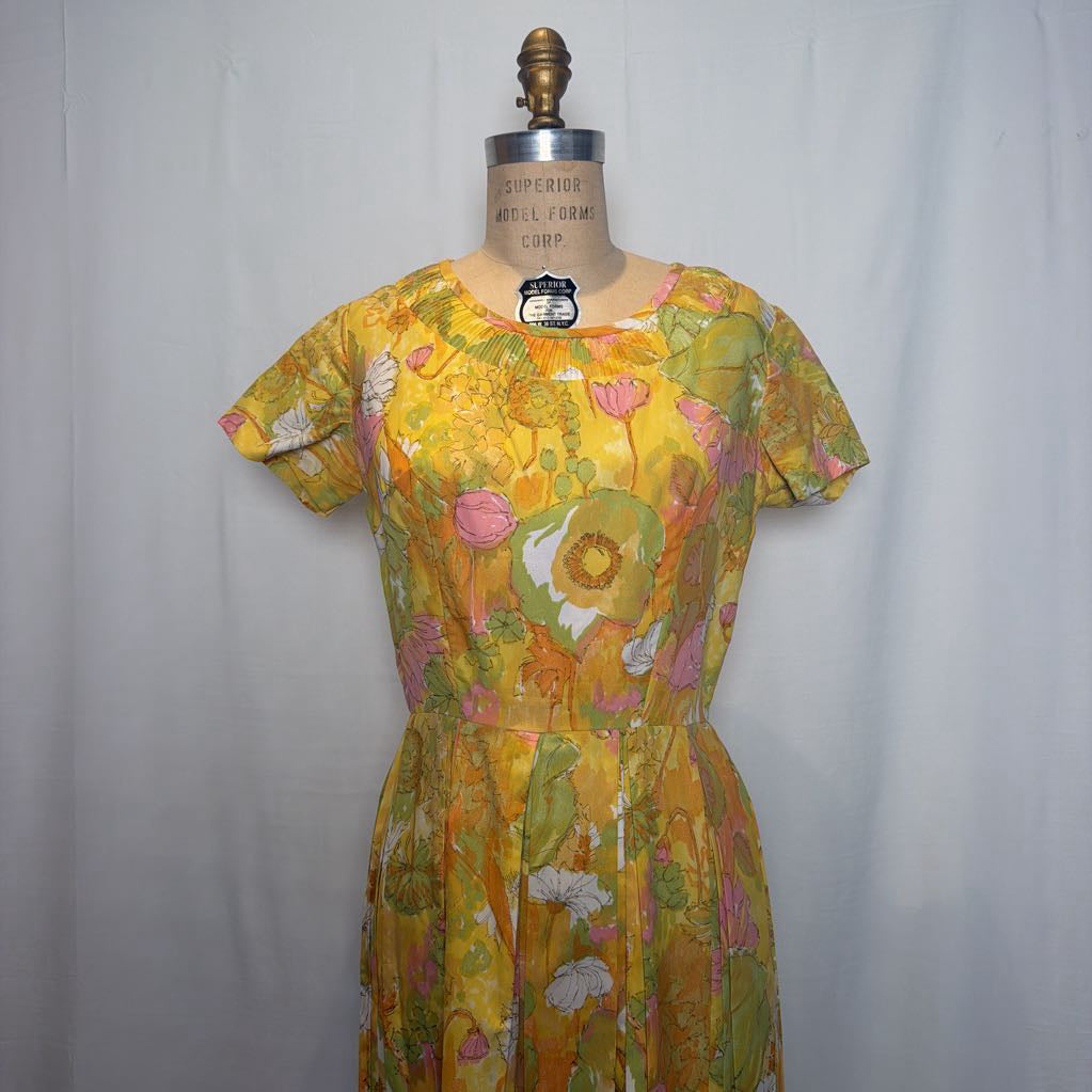 A vintage 1970s floral print dress displayed on a wooden dress form mannequin against a light gray fabric backdrop. The dress features a vibrant yellow base with pink, green, and orange botanical patterns.