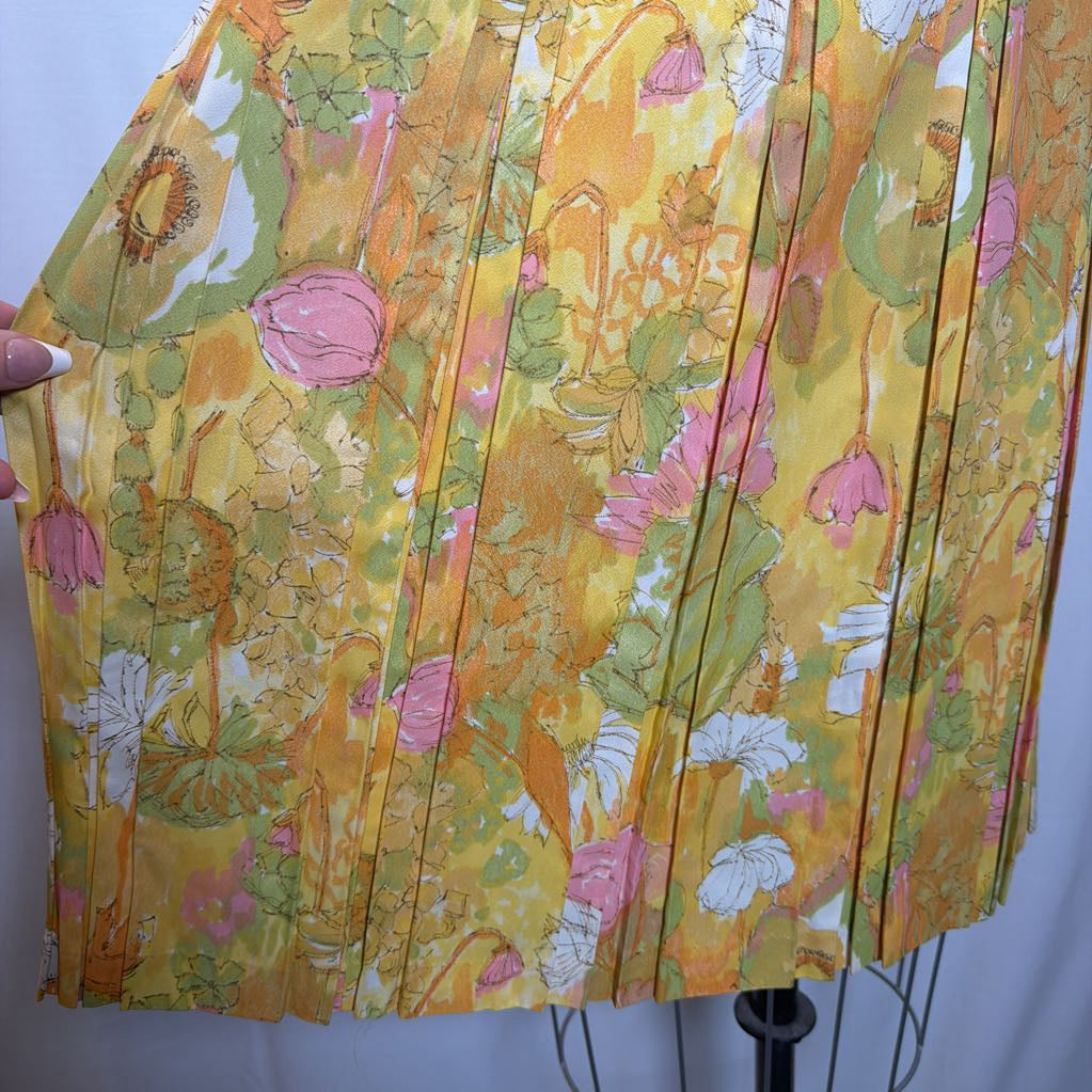A close-up of a pleated skirt or fabric panel with a watercolor-style floral print in yellow, pink, green, and white, held by a hand against a plain light background.
