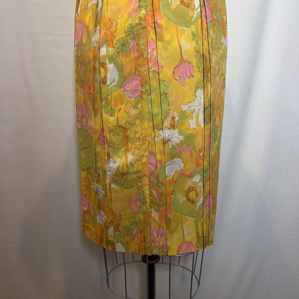 A pleated skirt displayed on a dress form, featuring a retro floral pattern in yellow, pink, green, and white, set against a neutral gray fabric backdrop.