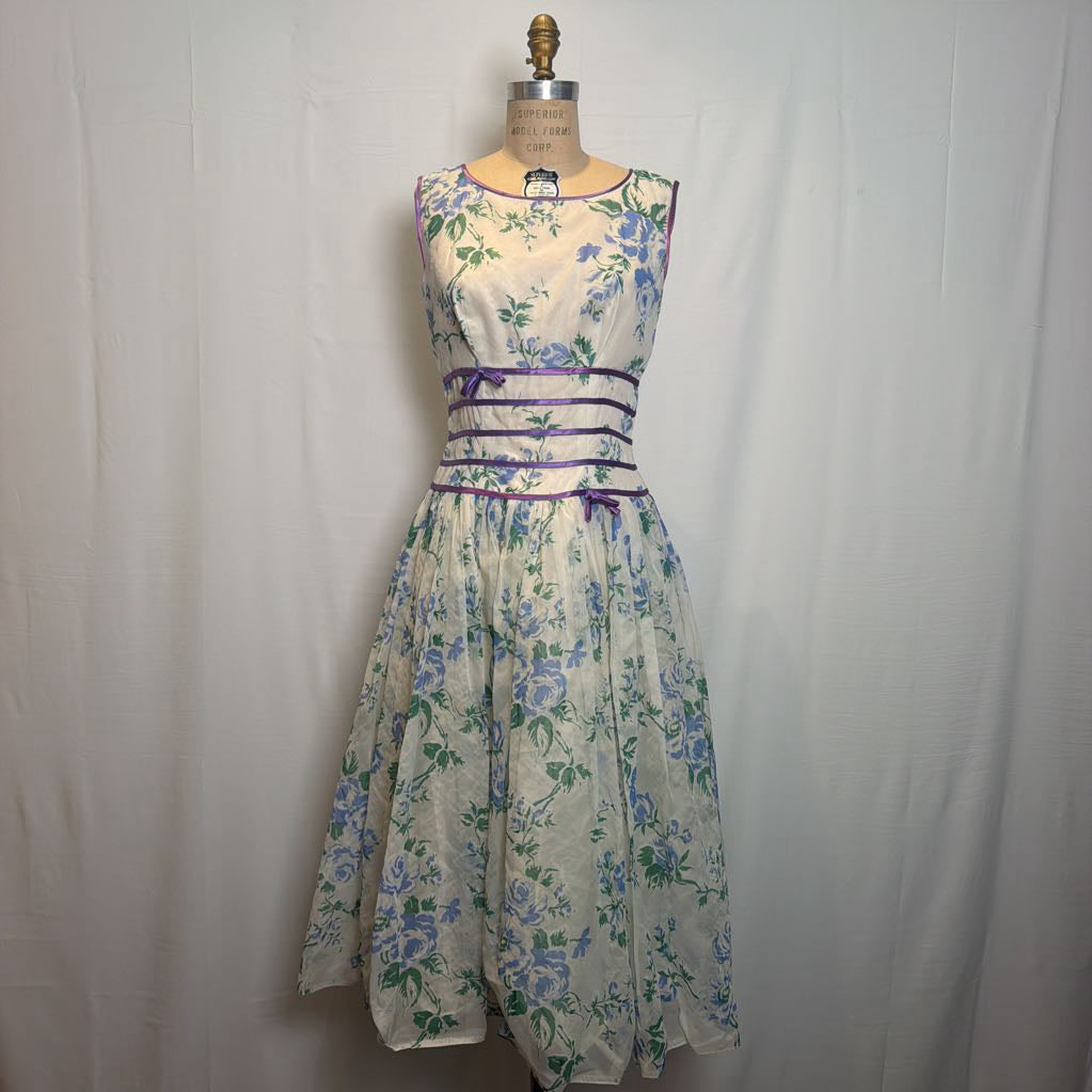 A sleeveless A-line dress displayed on a dress form against a white fabric backdrop. The dress has a cream base with a blue and green floral print and is accented with purple sash details at the waist.