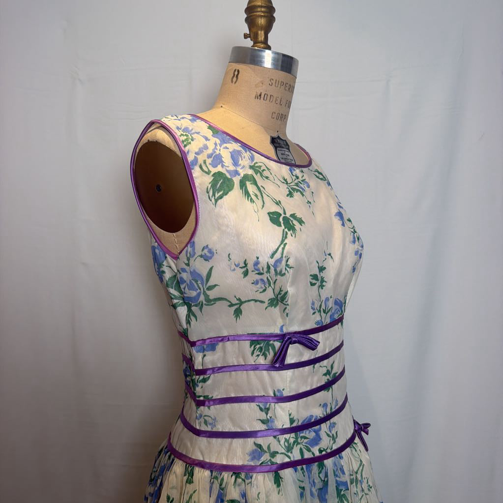 A sleeveless cream-colored dress with a blue and green floral print, featuring purple trim and waist ties, displayed on a vintage wooden dress form against a white fabric backdrop.