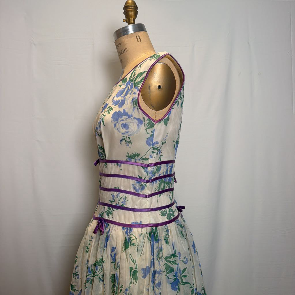 A sleeveless vintage-style dress with a blue and green floral print on a cream background, styled on a wooden dress form. The dress features purple ribbon accents at the waist and a purple trim around the neckline.