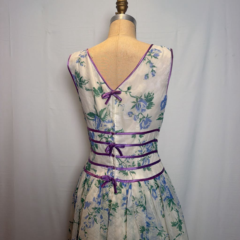 A sleeveless V-neck dress displayed on a dress form, featuring a cream-colored base with a blue and green floral print and purple trim details.