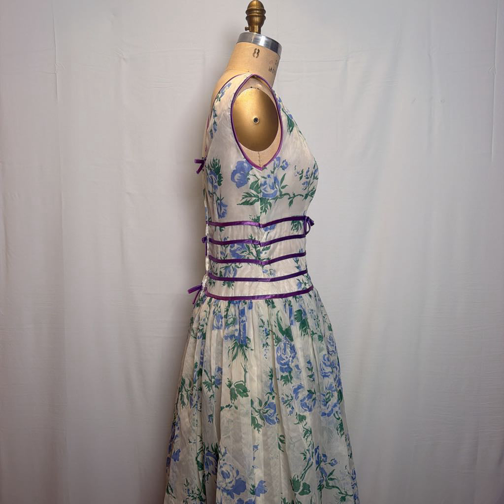 A sleeveless floral dress displayed on a dress form mannequin against a white fabric backdrop. The dress has a cream base with blue and green floral print and purple trim details.