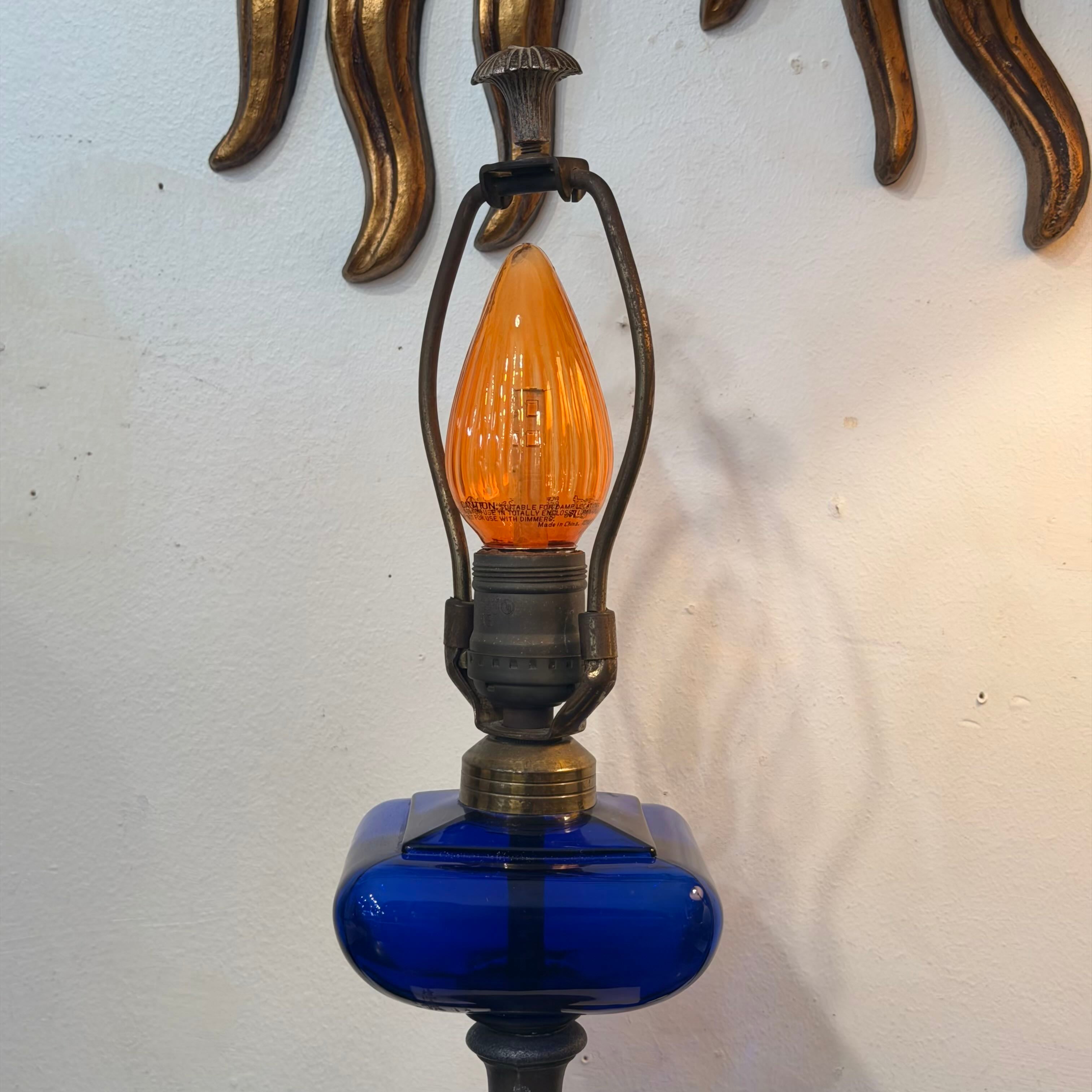 A vintage-style wall lamp featuring a cobalt blue glass base, a dark metal arched cage, and a ribbed orange glass bulb, shown against a white wall with a gold sunburst mirror decoration above.