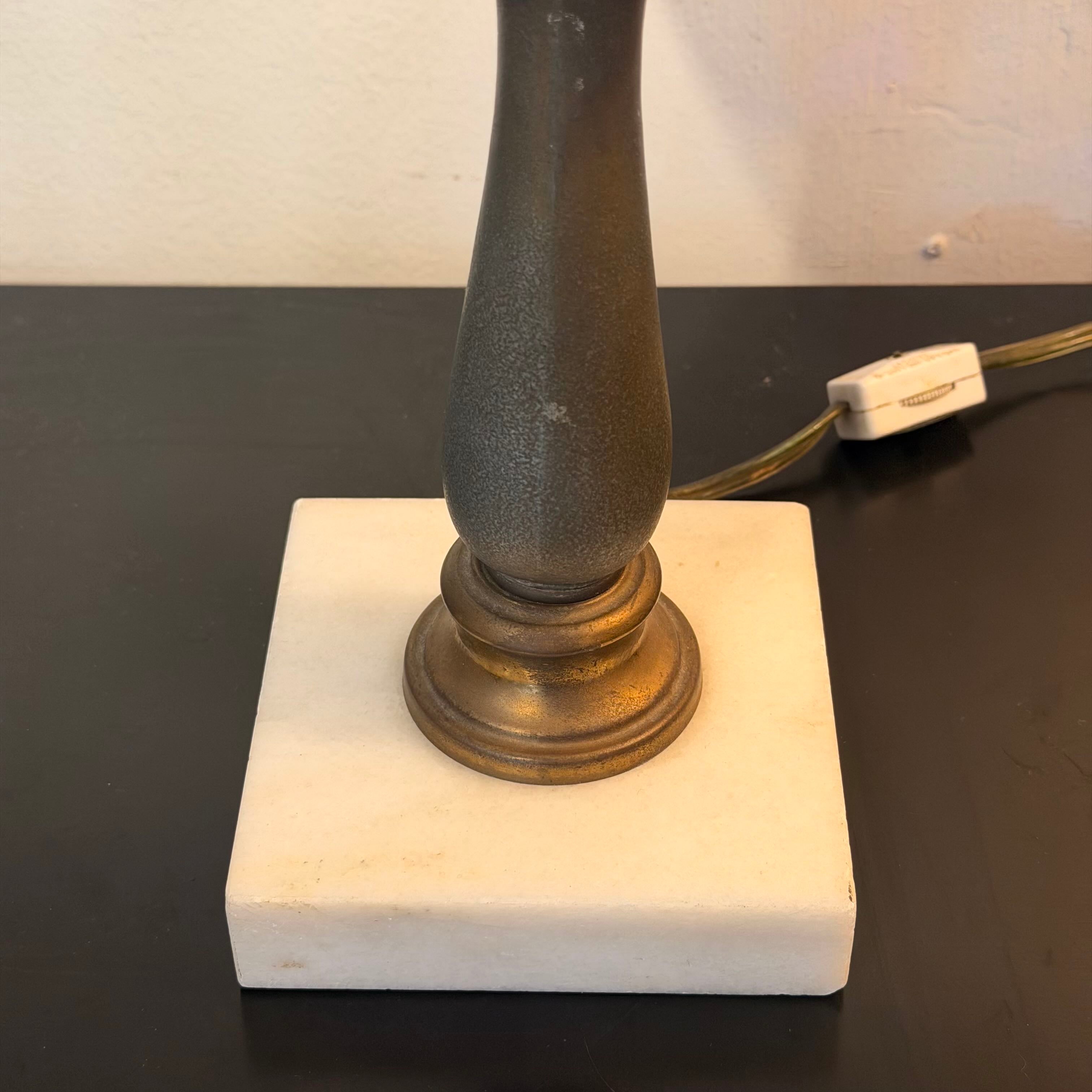 A close-up of a table lamp base featuring a square off-white marble or stone plinth, a textured dark gray or bronze-toned central column, and a gold-toned circular collar, with a black power cord running to the right on a dark tabletop.