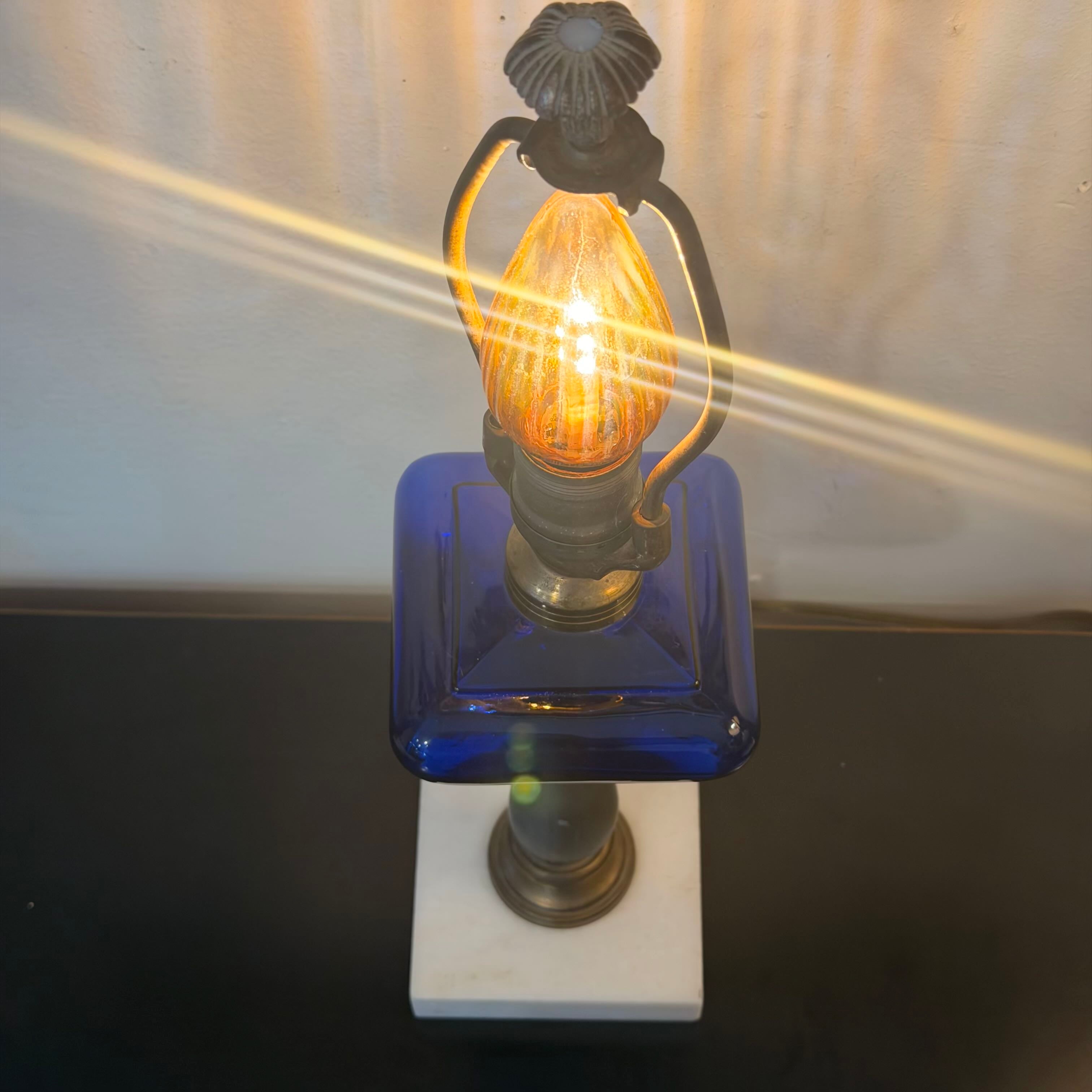 A vintage-style table lamp with a glowing amber filament bulb, a dark metal lantern-shaped top, a cobalt blue glass square body, and a white rectangular base, photographed against a light wall with strong light rays.
