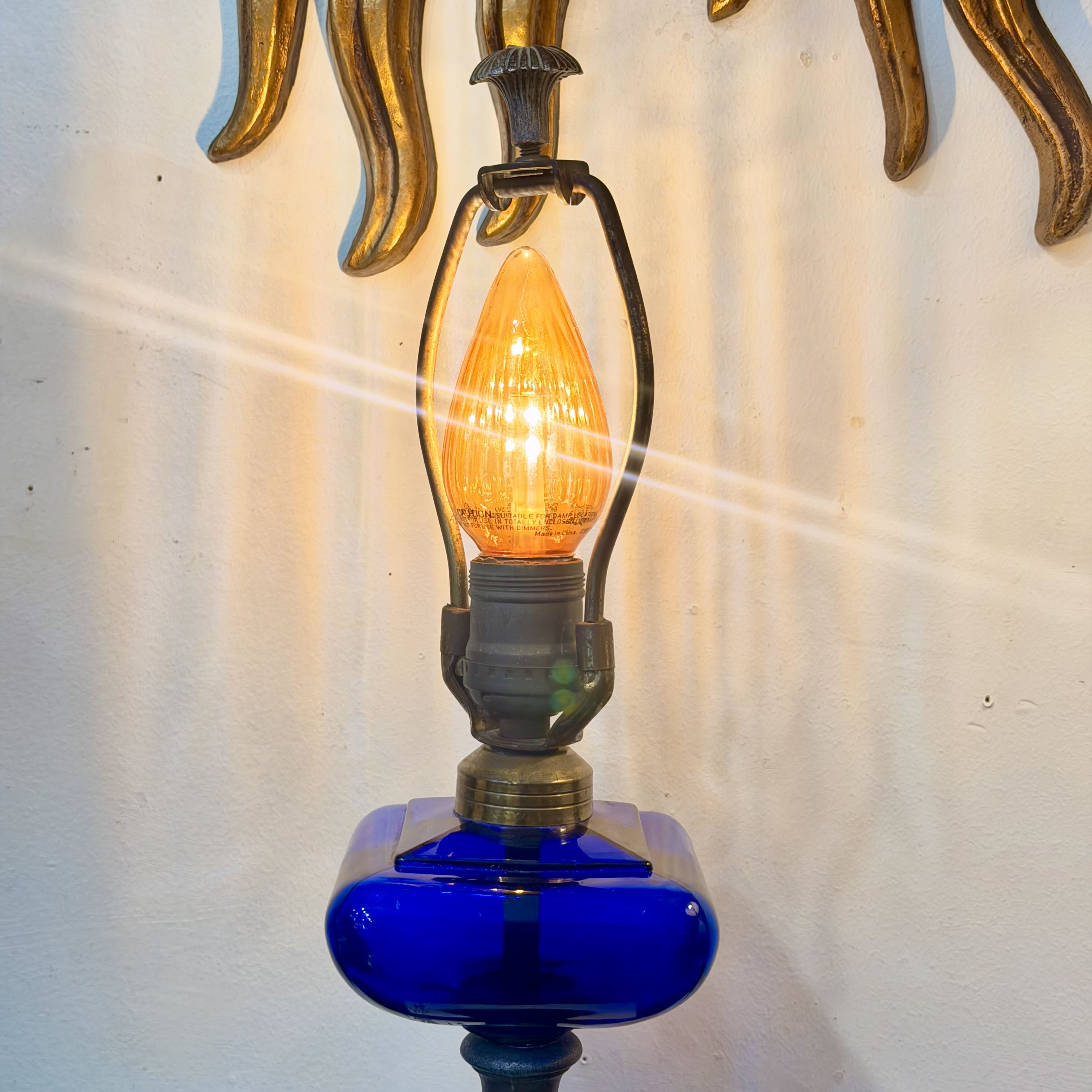 A close-up of a vintage-style wall lamp with a glowing amber bulb inside a black metal frame, mounted on a cobalt blue glass base against a white wall with a gold decorative element above.