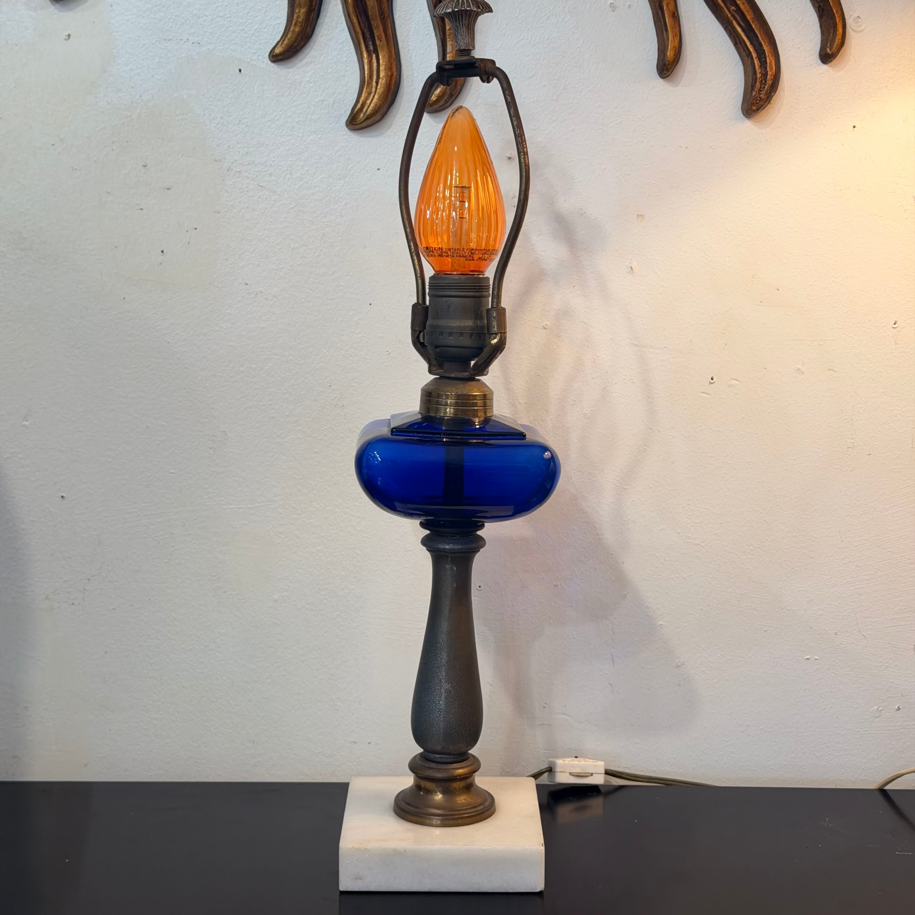 A vintage-style table lamp with a cobalt blue glass bulb holder, a dark metal shade and base, and an illuminated orange bulb, set on a white marble base against a white wall.