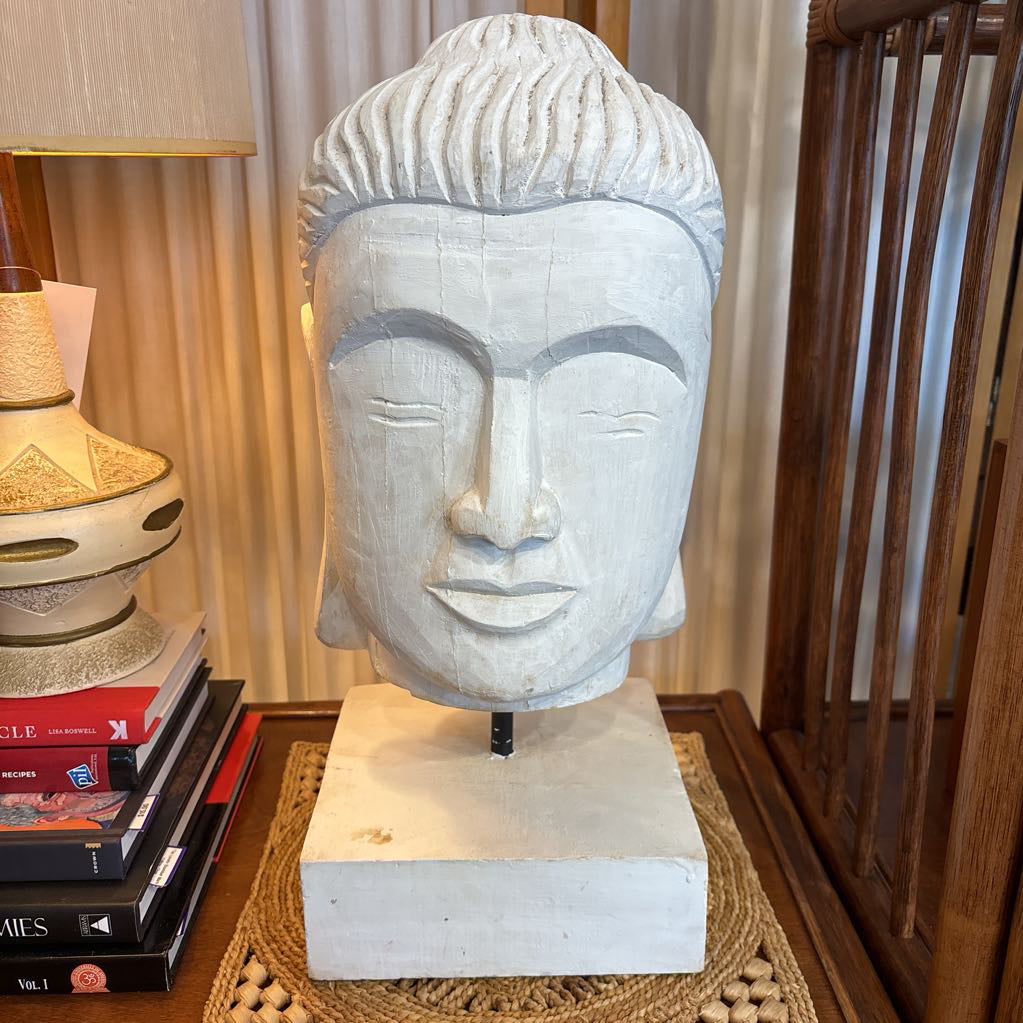 A large white carved Buddha head sculpture on a matching rectangular base, displayed on a wooden table in a home interior setting next to a lamp and books.