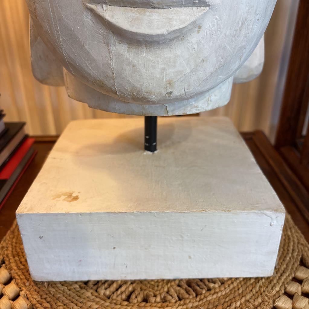 A close-up of a carved stone or wood sculpture base mounted on a square pedestal, with a black support rod extending upward into a large pale face. The sculpture sits on a woven round mat on a wooden table, with books and curtains in the background.