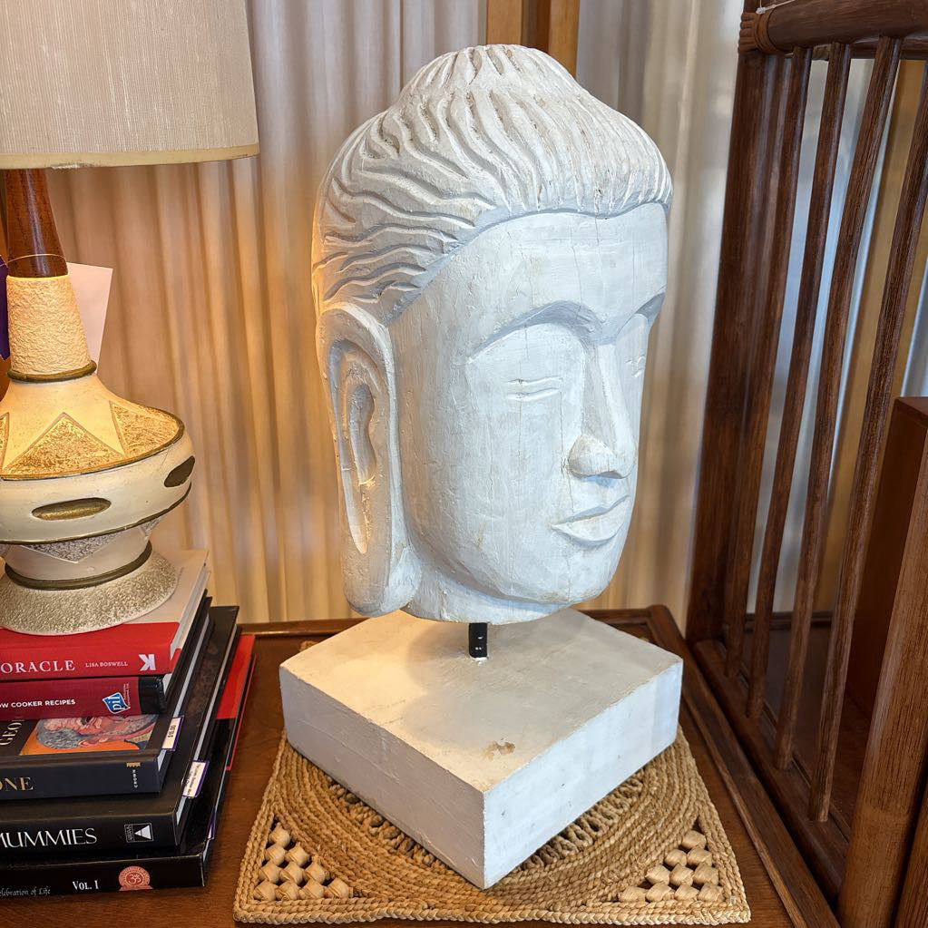 A large carved Buddha head sculpture in a light gray stone finish, mounted on a matching square base, displayed on a woven placemat on a wooden table next to a lamp and books.