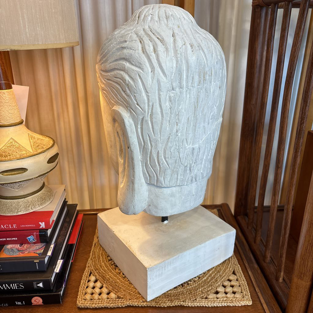 A carved stone Buddha head sculpture displayed on a square pedestal base, set on a woven placemat on a wooden table in a home interior next to a lamp and books.