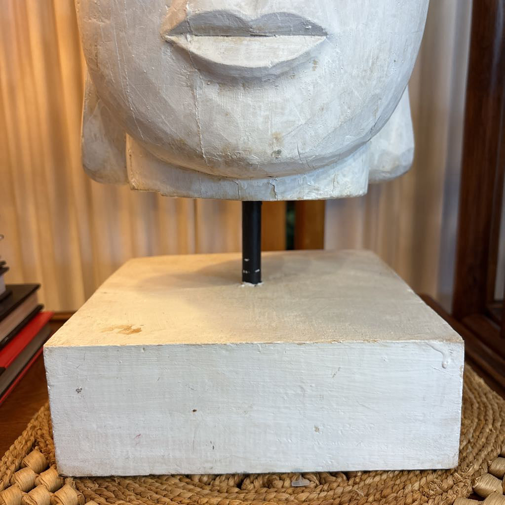 A carved wooden Buddha head sculpture mounted on a square white-painted base with a black central support rod, photographed indoors on a woven mat.