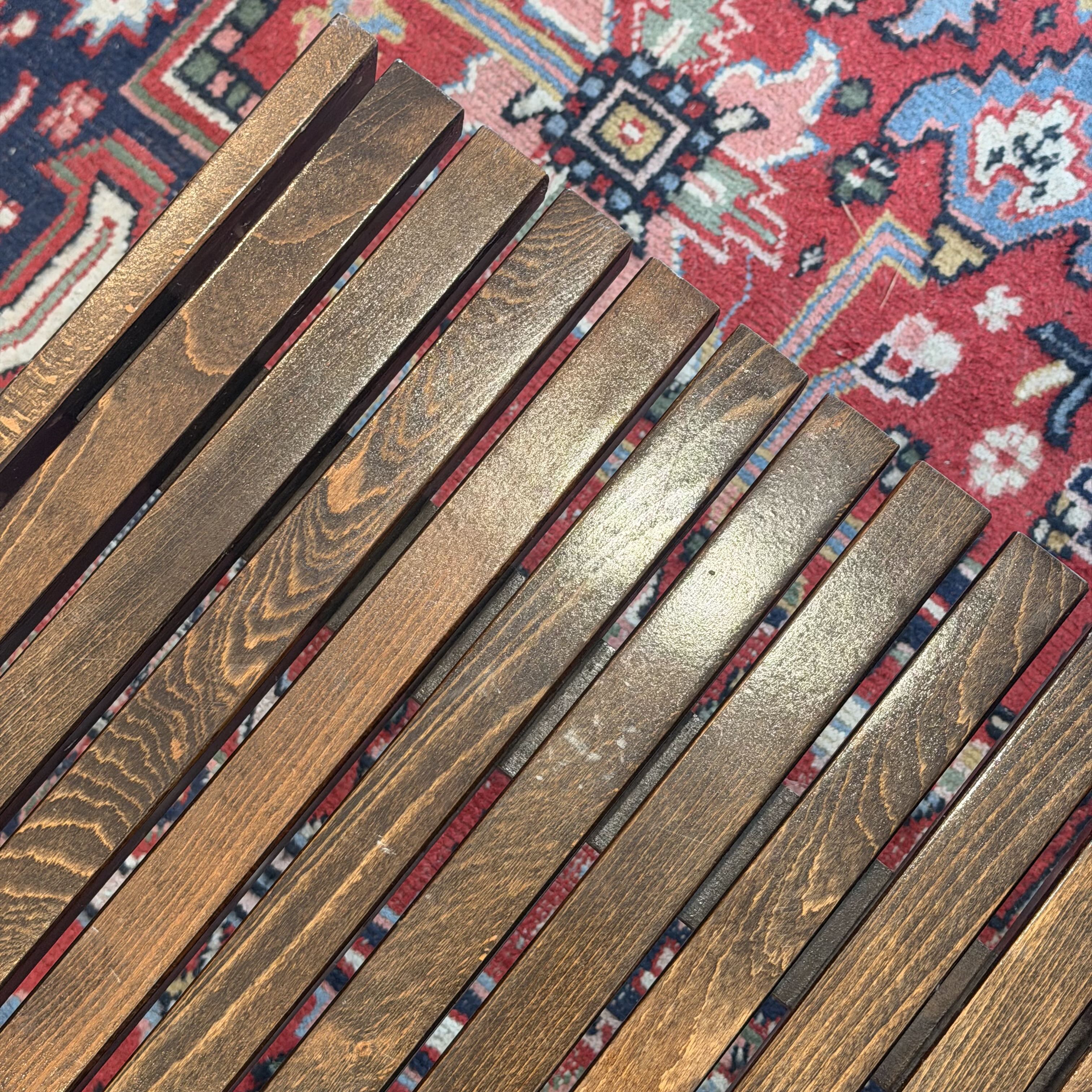 Close-up of a wooden slatted surface, likely a chair or bench, with a red patterned rug visible underneath in the background.