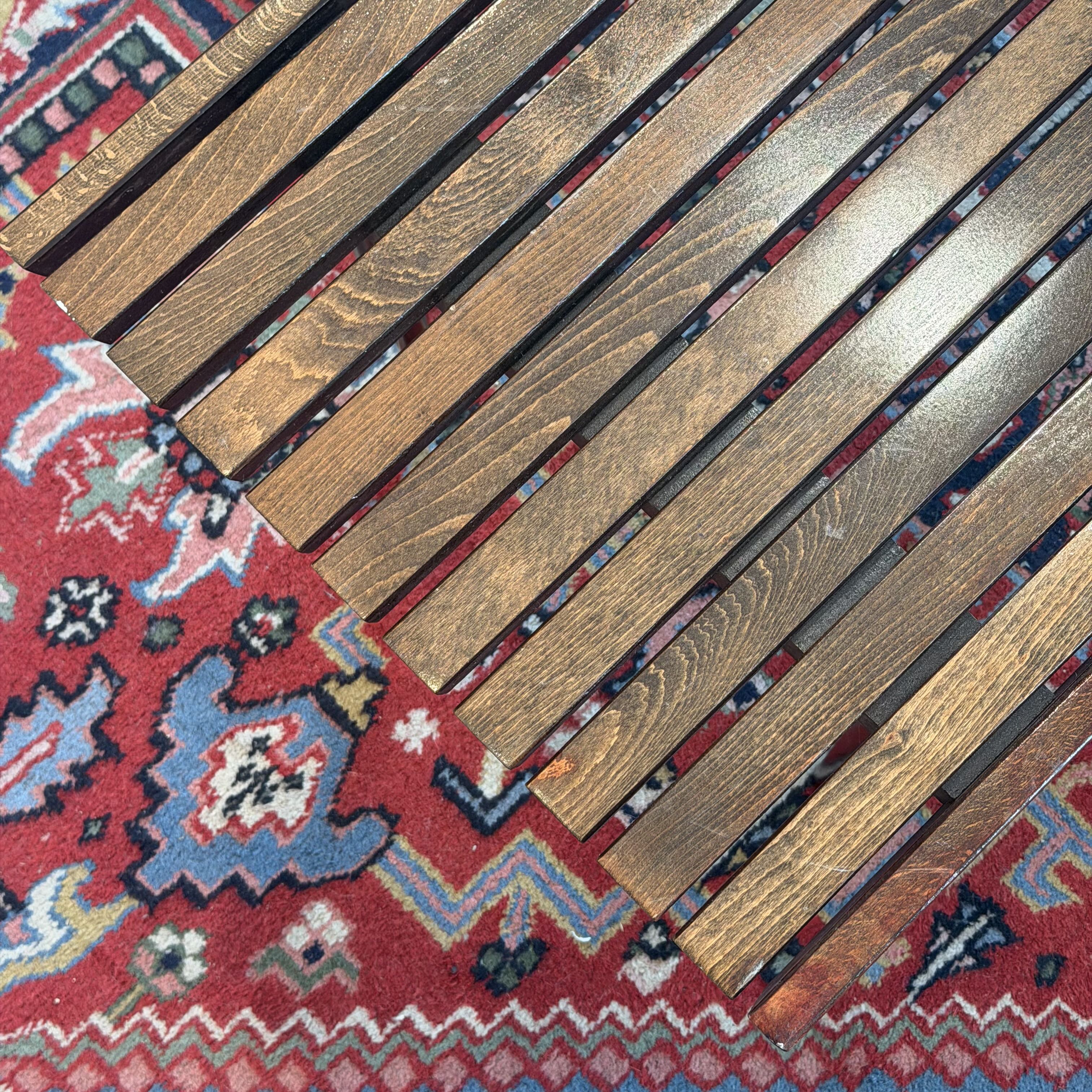 A close-up of a wooden slat surface, likely a table or bench, laid diagonally over a red and blue patterned rug.