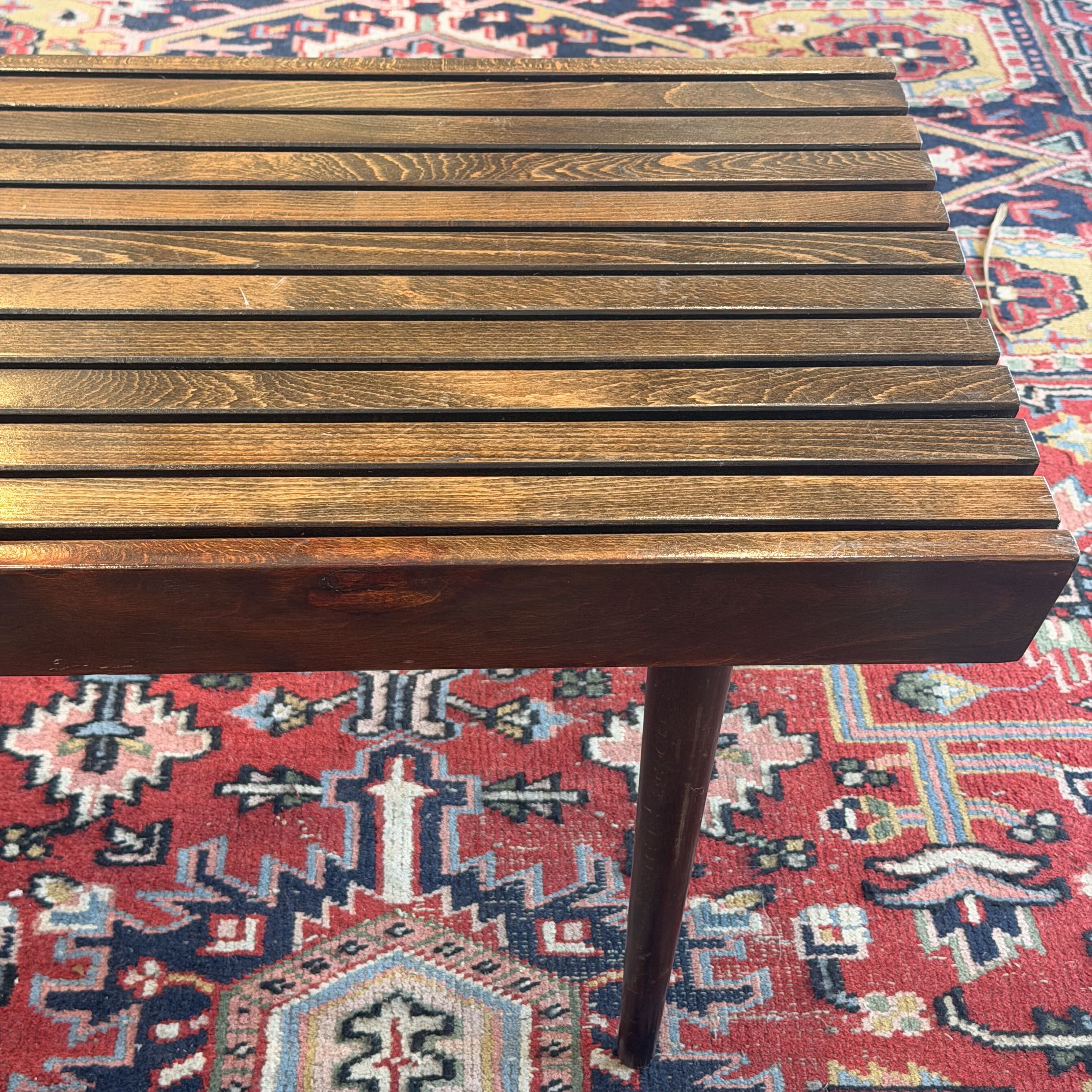 A mid-century modern wooden slat side table with a dark brown finish, shown from a high angle resting on a red and blue patterned oriental rug.