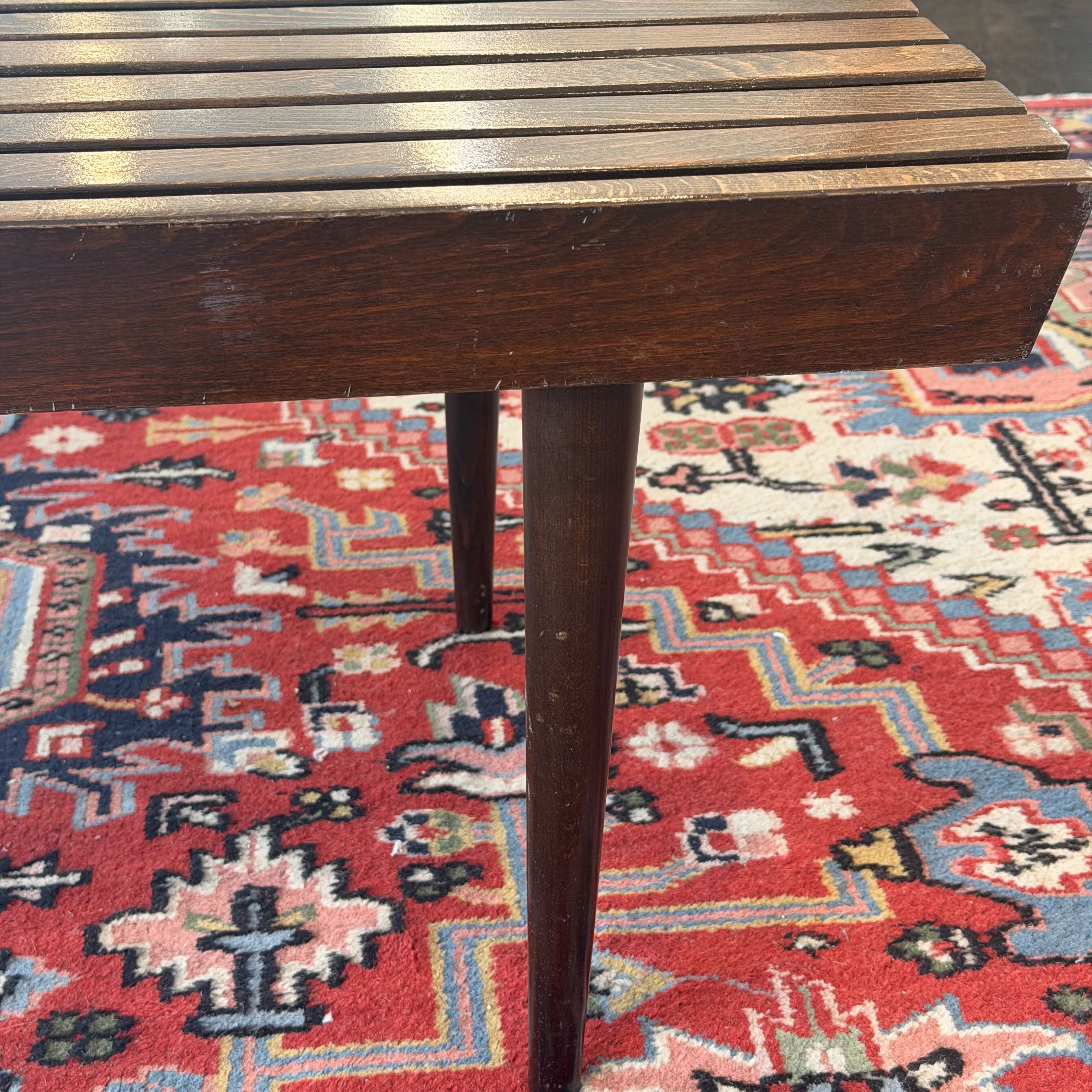 Close-up of a dark brown wooden slat bench or stool leg resting on a red and blue patterned oriental rug.