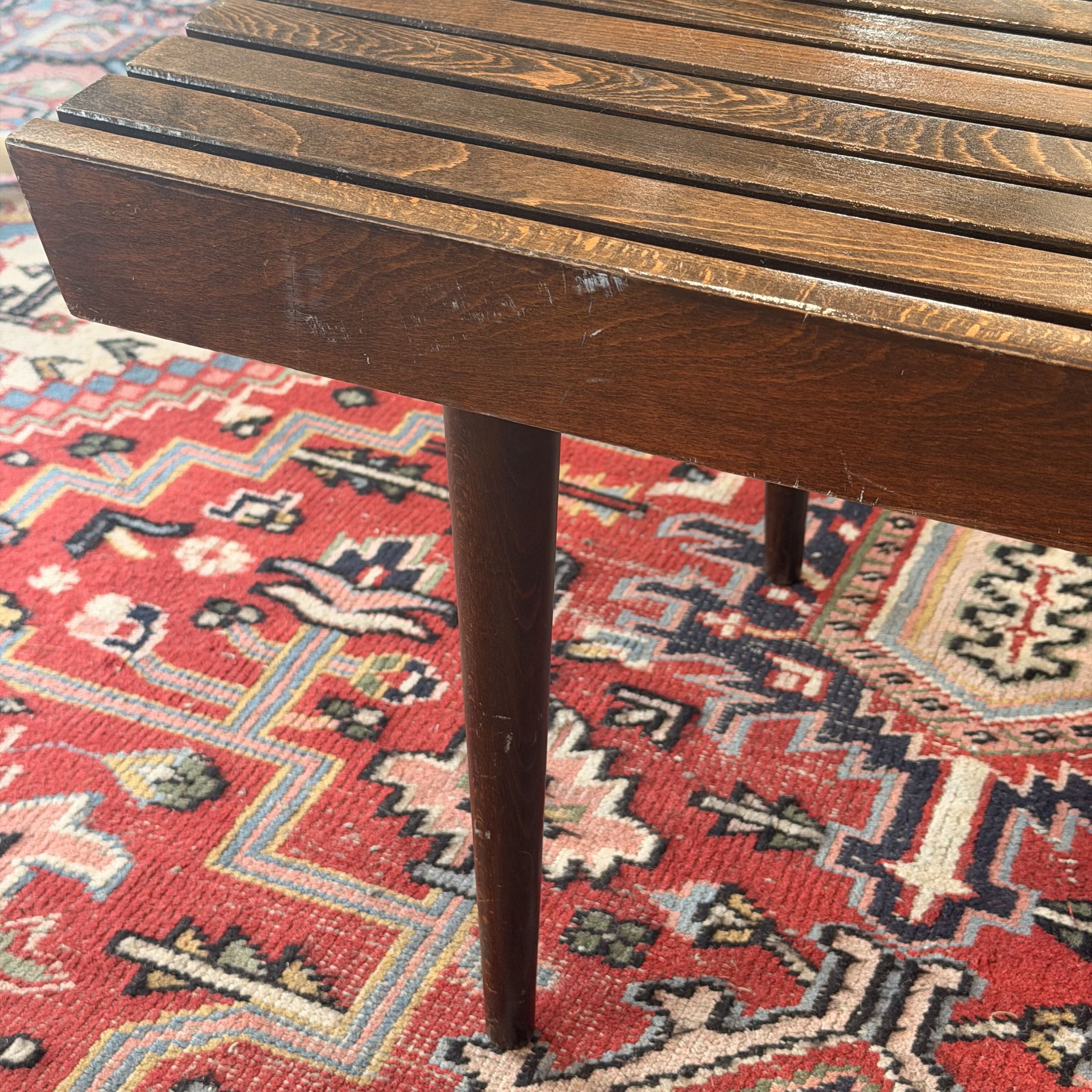 A close-up of a dark brown wooden slat bench or stool leg resting on a red patterned oriental rug.
