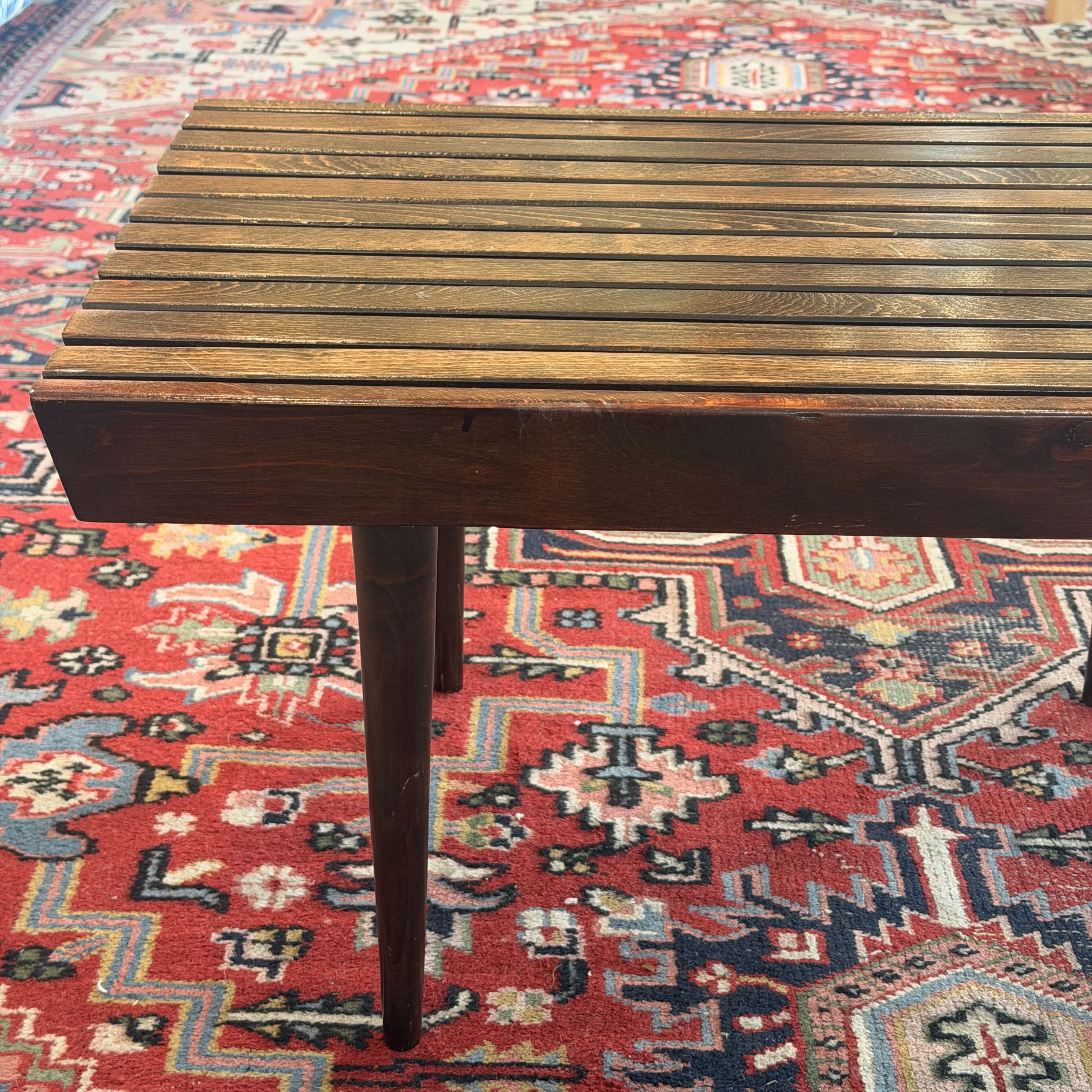A dark brown wooden slat bench or stool with a single leg, positioned on a red and blue patterned oriental rug.