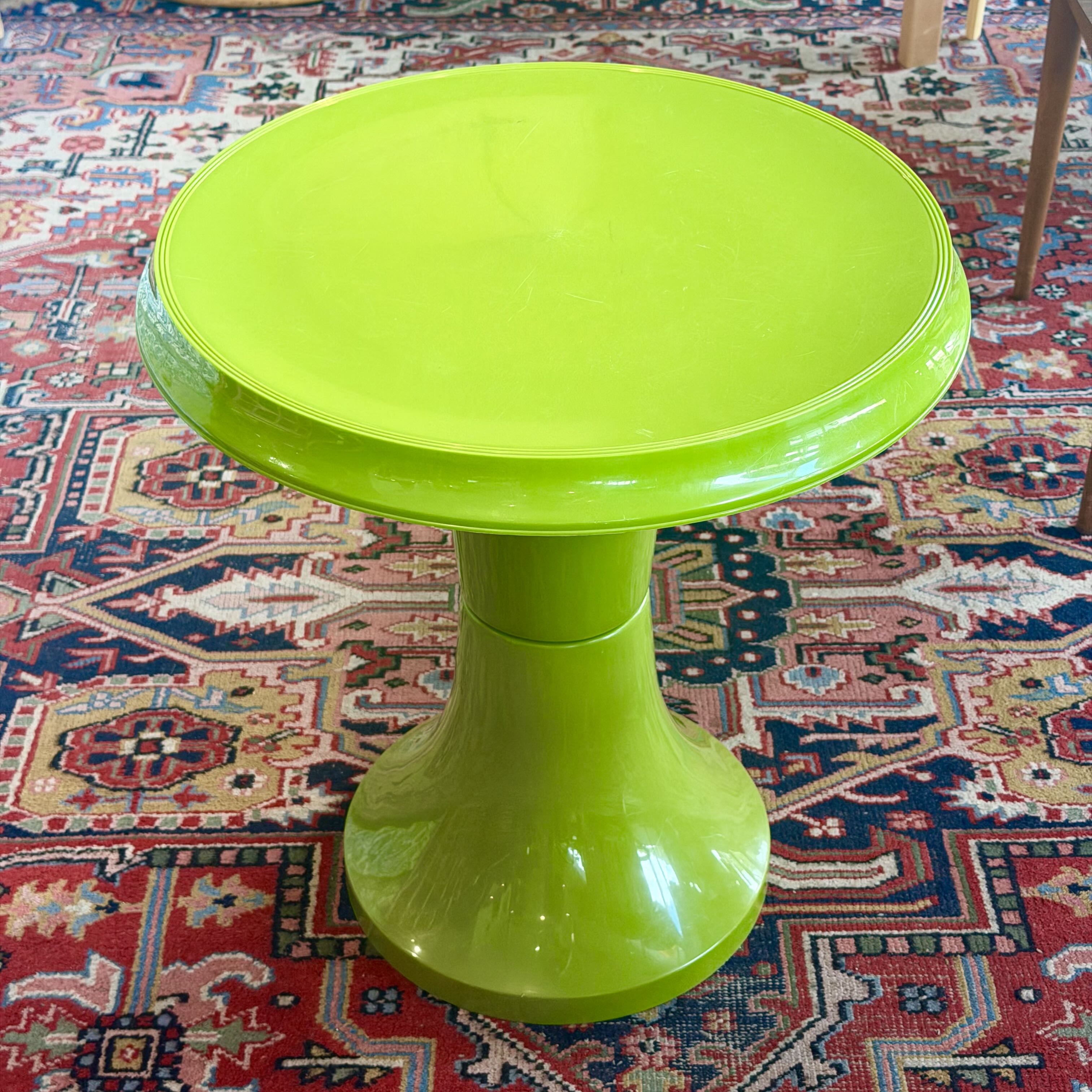 A bright lime green pedestal side table with a glossy finish, set on a patterned red and blue oriental rug.