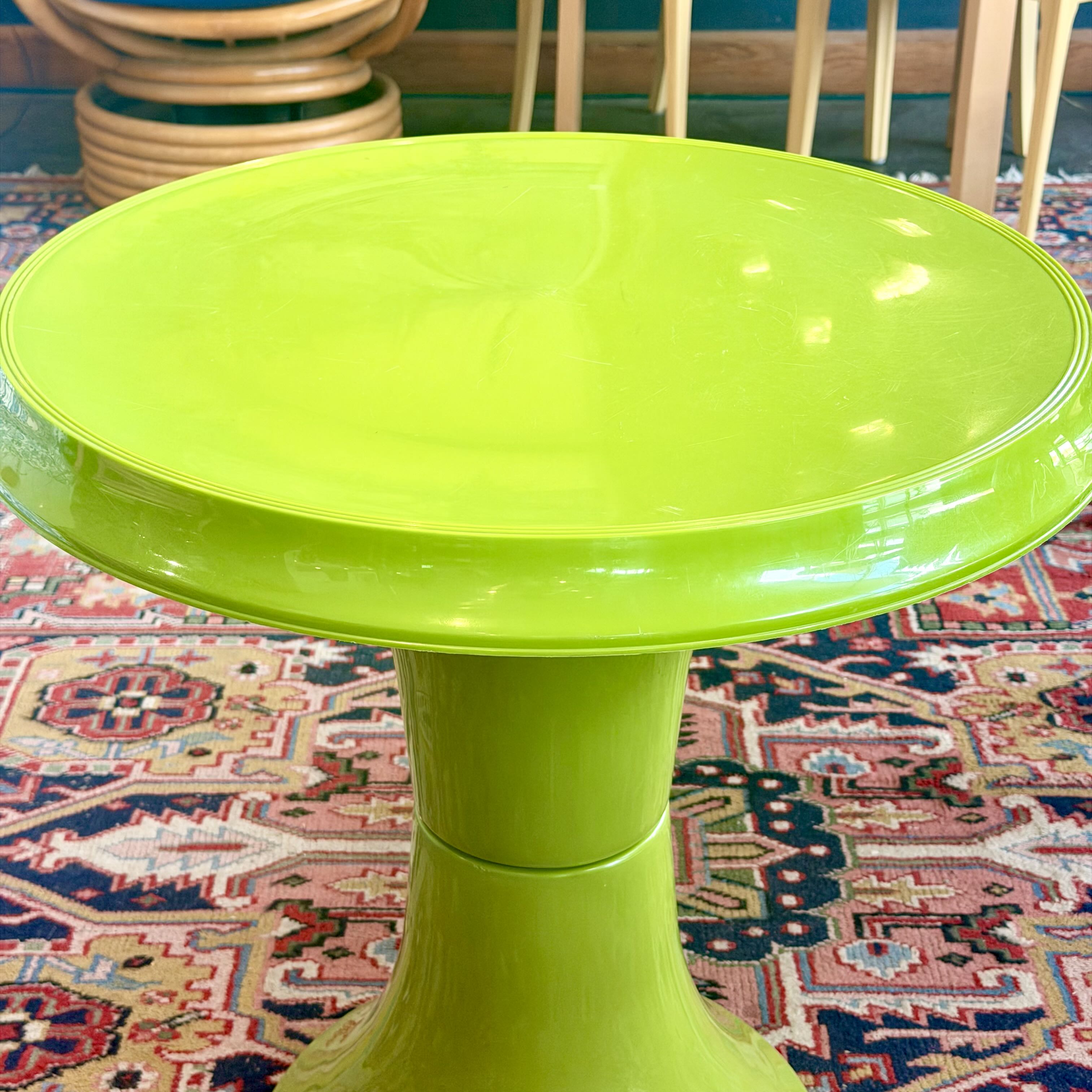 A bright lime green pedestal side table with a glossy finish, set on a patterned rug with wooden chairs in the background.