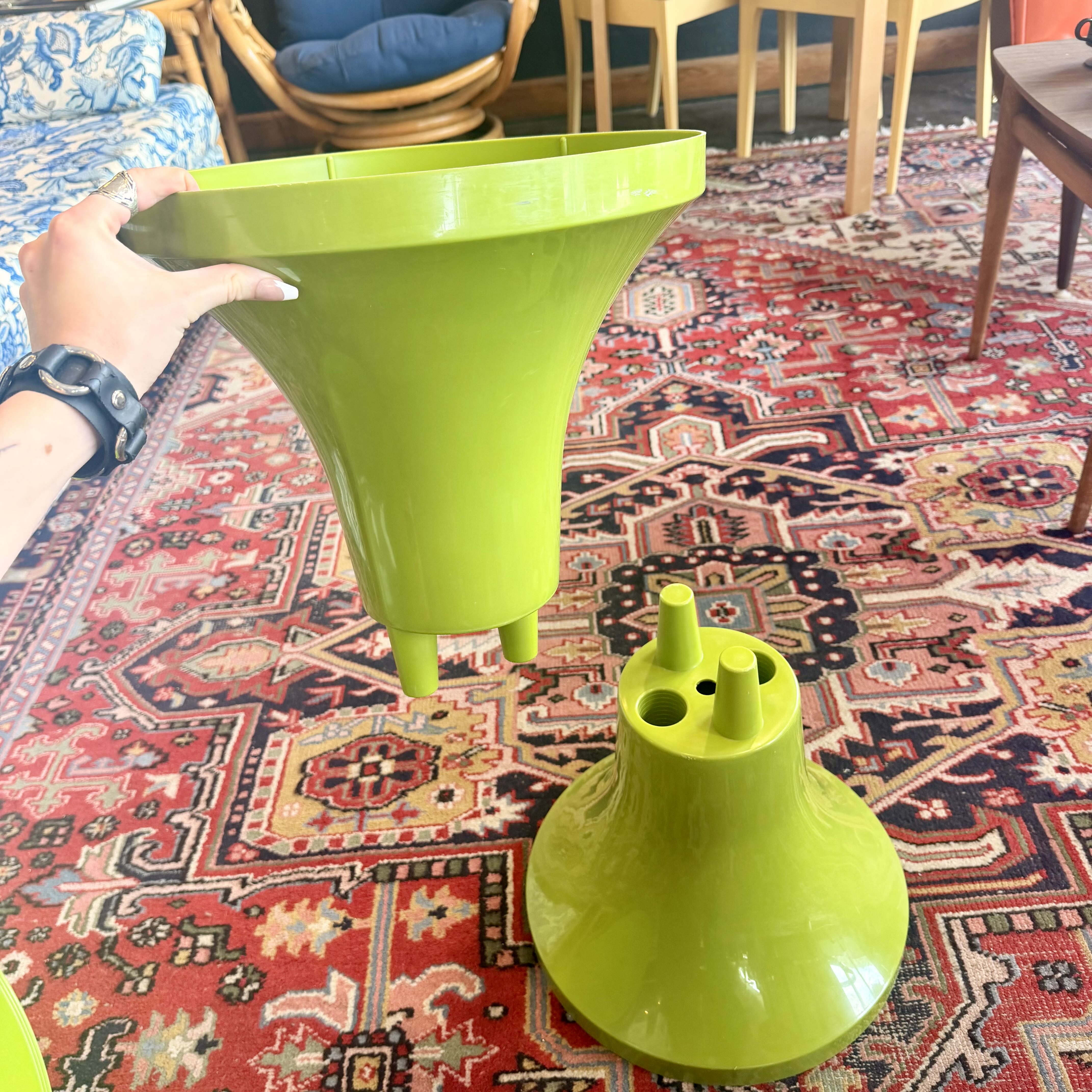 A bright lime green plastic two-piece set, likely a small table base or decorative stand, being held by a hand over a red patterned oriental rug in a living room setting.