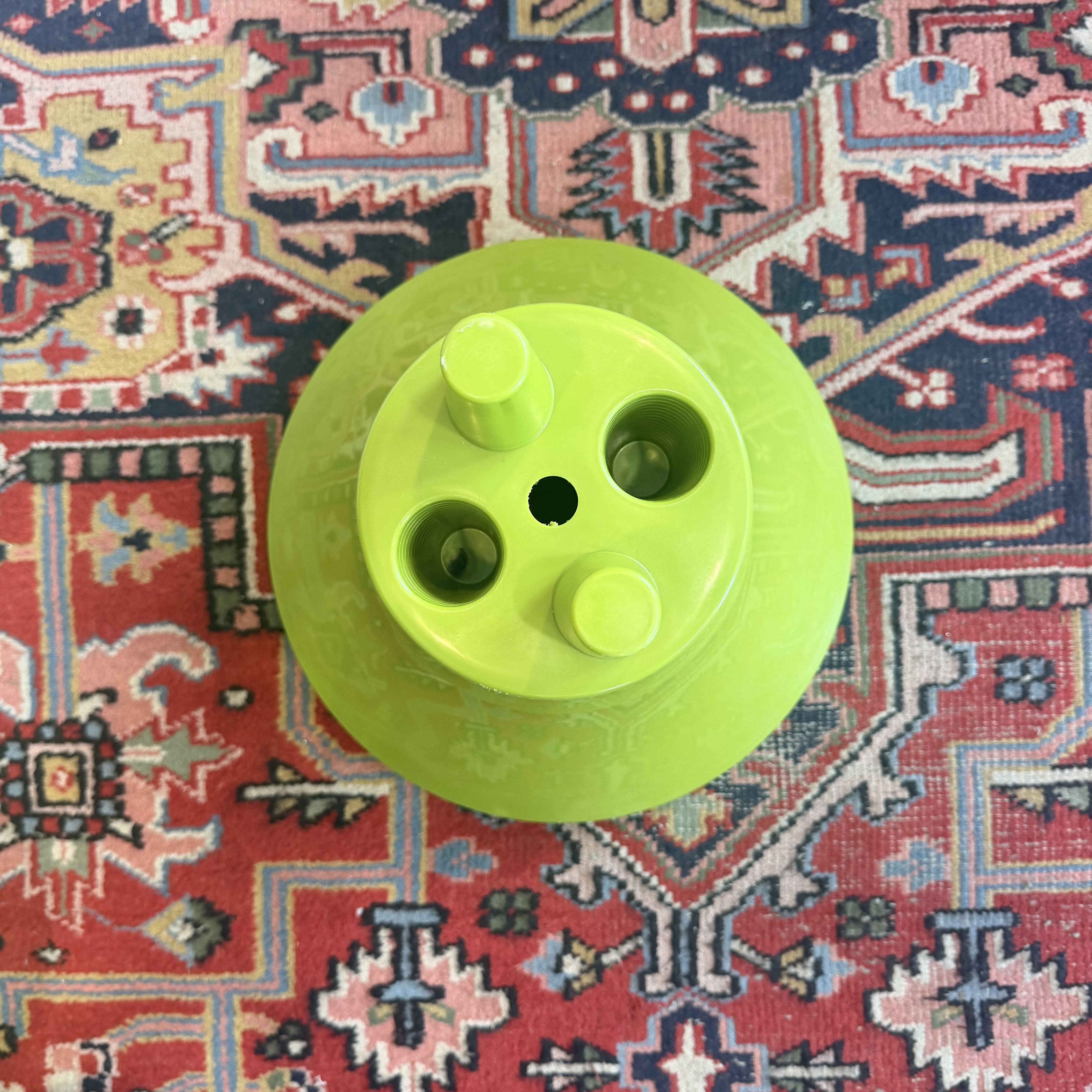 A bright lime green plastic circular component with three cylindrical protrusions and three circular openings, photographed from above on a patterned red and blue rug.