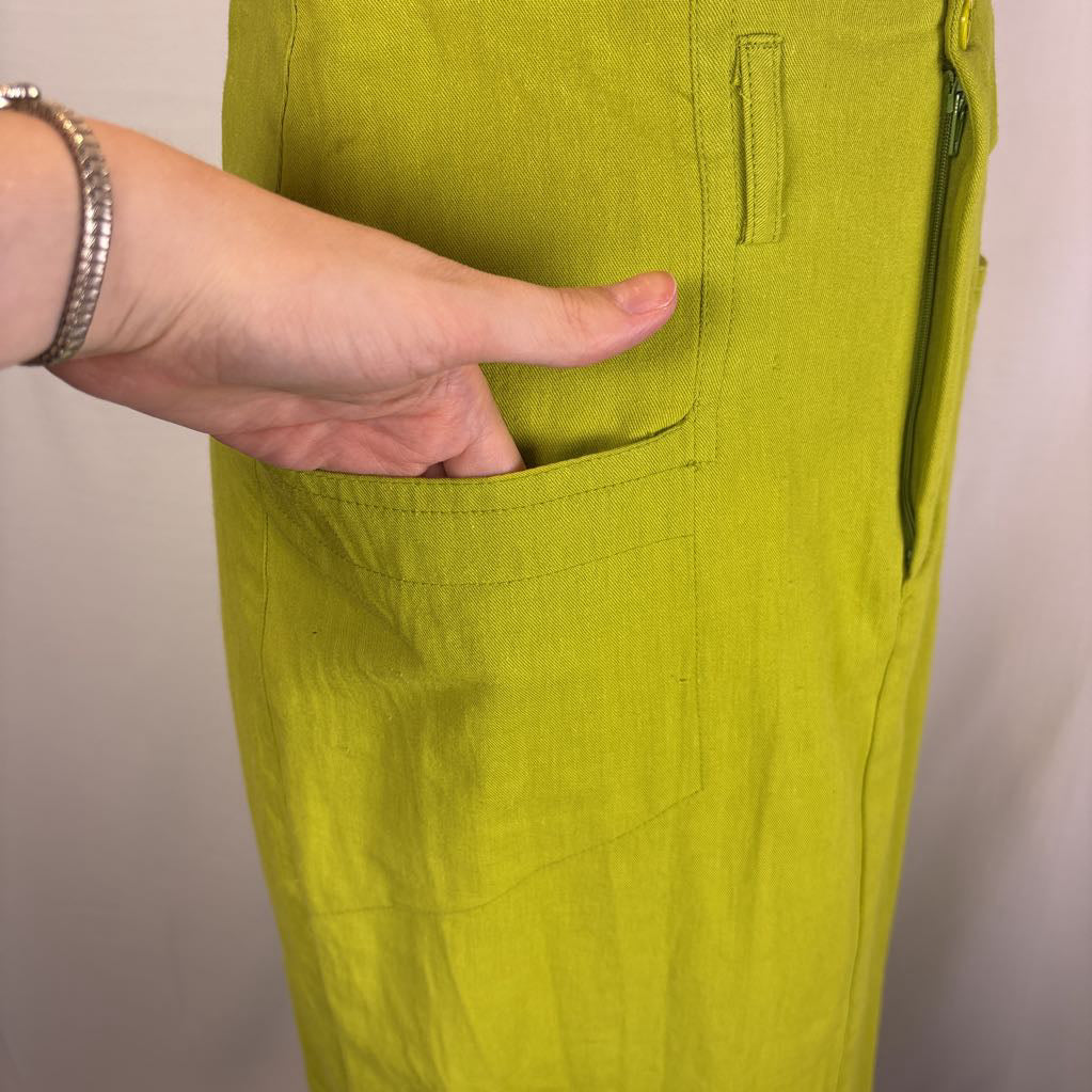 A close-up of a person's hand reaching into a front pocket of bright yellow-green linen trousers, showing the fabric texture and pocket detail.
