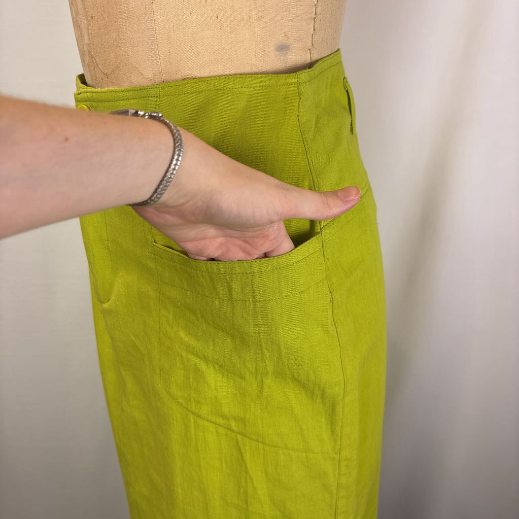 A close-up of a person's hand reaching into a front pocket of a bright yellow-green linen apron or skirt, shown on a dress form against a plain light background.