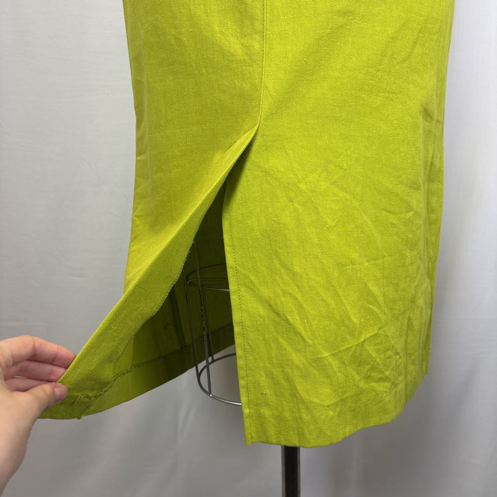 A close-up of a bright yellow-green skirt or drape on a dress form, with a hand pulling the lower left side to show a slit or hem detail against a plain light background.