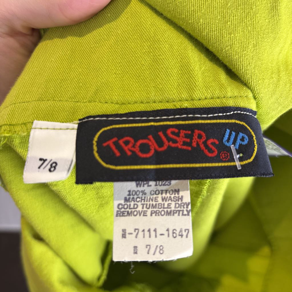 Close-up of the inside neck label of a bright yellow-green garment. The black brand label reads 'TROUSERS UP' in red and blue, with a white size tag showing '7/8' and a cotton care tag below it.