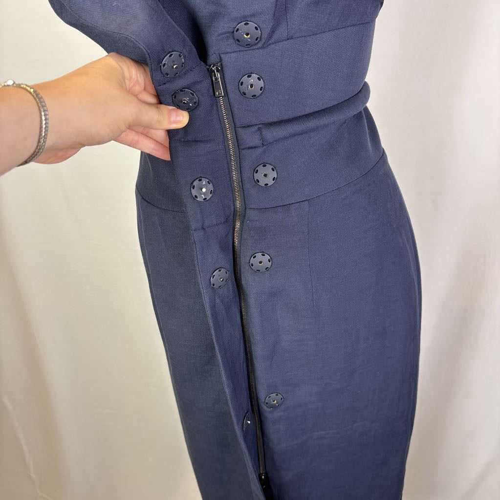 Close-up of a navy blue dress with a front zipper and decorative buttons, being held at the waist by a hand against a light fabric backdrop.