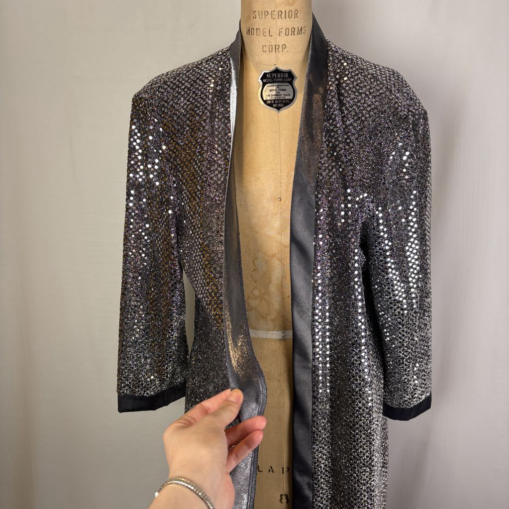 A black and silver sequined open-front jacket displayed on a vintage dress form, with a hand holding the left side to show the fabric movement.