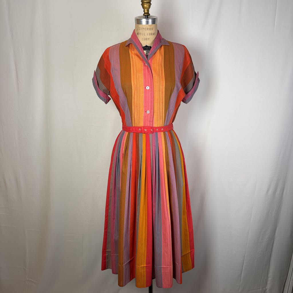 A short-sleeve collared dress displayed on a dress form against a white fabric backdrop. The dress features vertical stripes in orange, pink, red, yellow, and purple, with a matching waist belt.