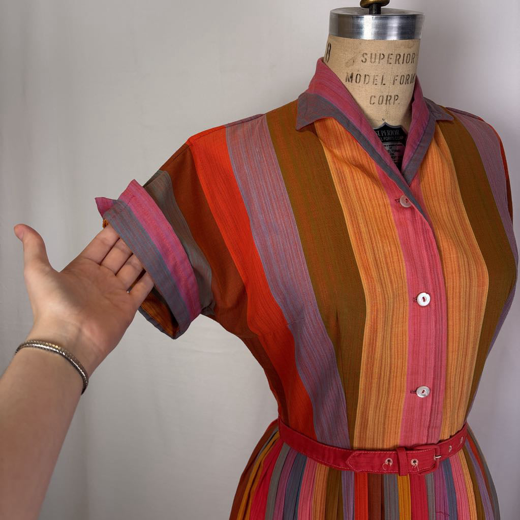 A vintage striped button-up shirt dress displayed on a wooden mannequin, with a person's hand adjusting the sleeve. The dress features vertical stripes in orange, red, pink, brown, and gray, with a red belt at the waist.