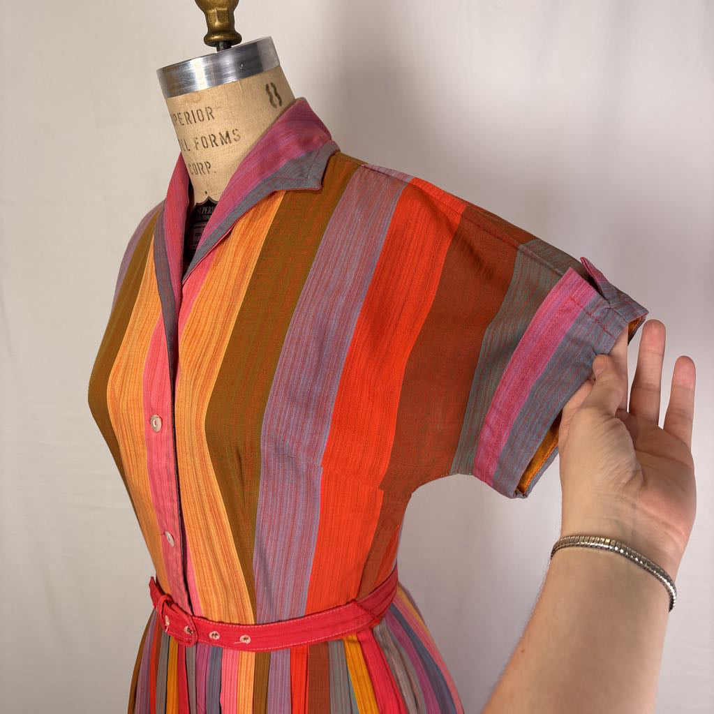 A multicolor vertically striped short-sleeve shirt dress displayed on a vintage dress form, with a hand adjusting the sleeve.