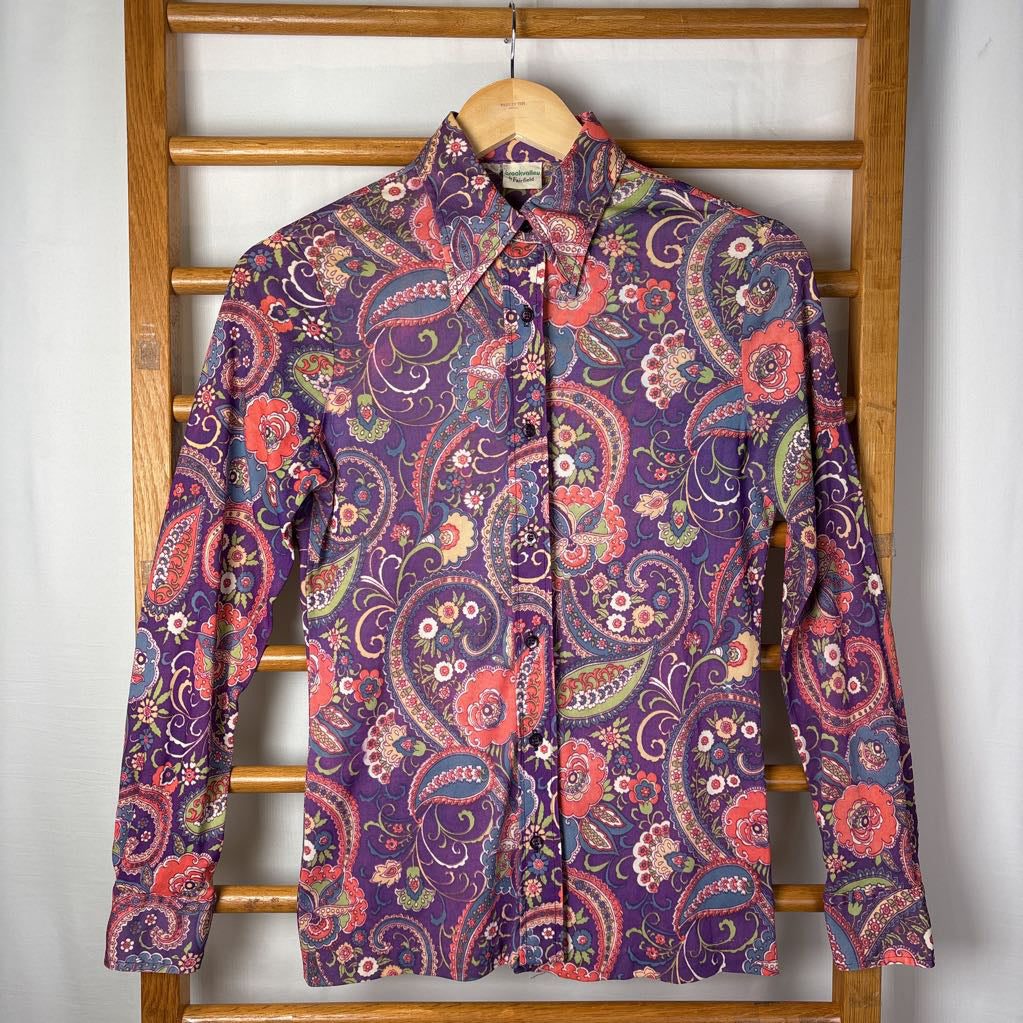 A vintage purple and paisley print button up long sleeve shirt on a hanger on a wooden rack. 