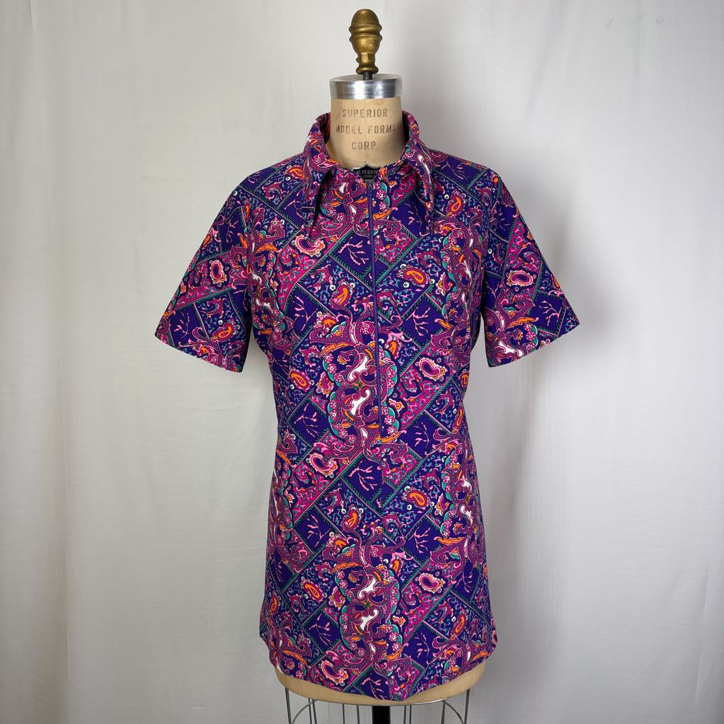 A short-sleeve button-up shirt with a vibrant purple, pink, and orange paisley pattern, displayed on a vintage wooden dress form mannequin against a white fabric backdrop.