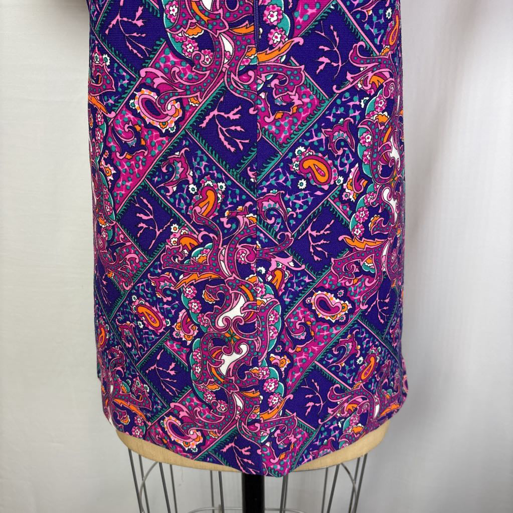 A close-up of a purple fabric with a vibrant paisley and diamond pattern in pink, orange, teal, and white, displayed on a dress form.