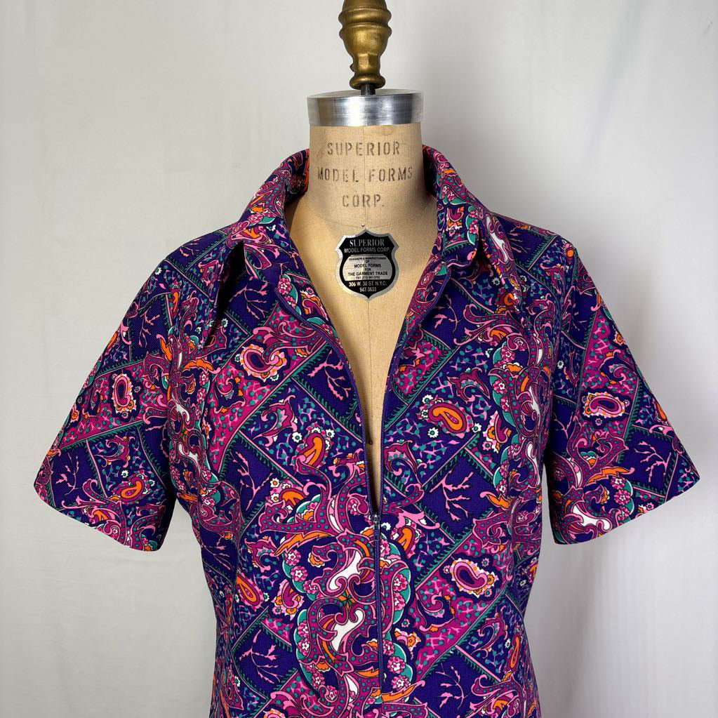 A short-sleeve button-front blouse with a bold paisley and geometric print in purple, pink, orange, and teal, displayed on a vintage dress form mannequin against a white fabric backdrop.