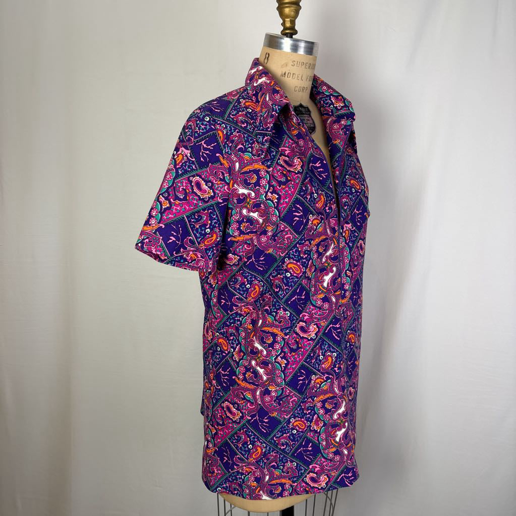 A short-sleeve button-up shirt with a vibrant purple, pink, and orange paisley pattern, displayed on a dress form mannequin against a neutral light gray fabric backdrop.