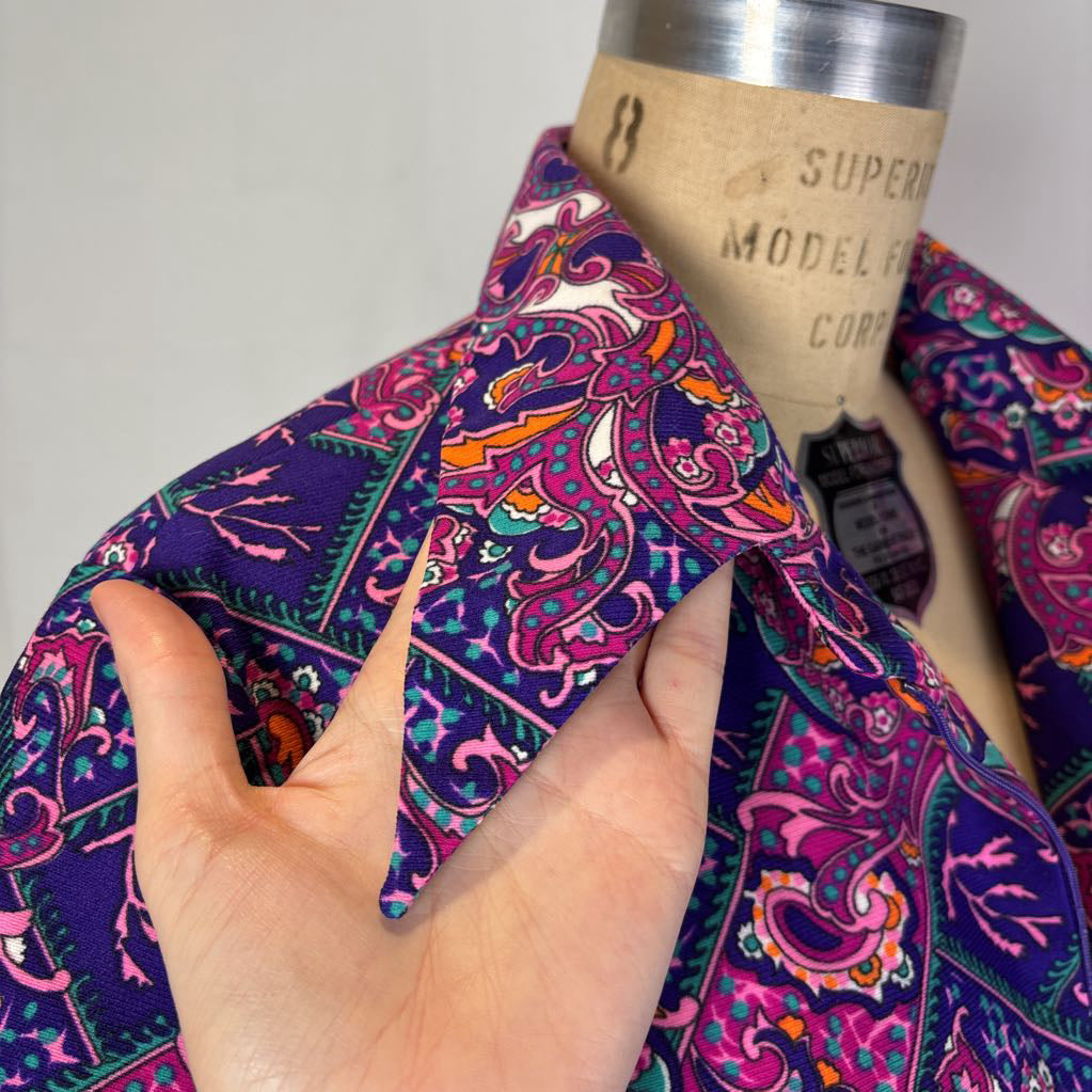 A close-up of a patterned collared shirt on a dress form, with a hand holding the collar to show its shape and fabric. The shirt features a vibrant purple, pink, orange, and teal paisley pattern.