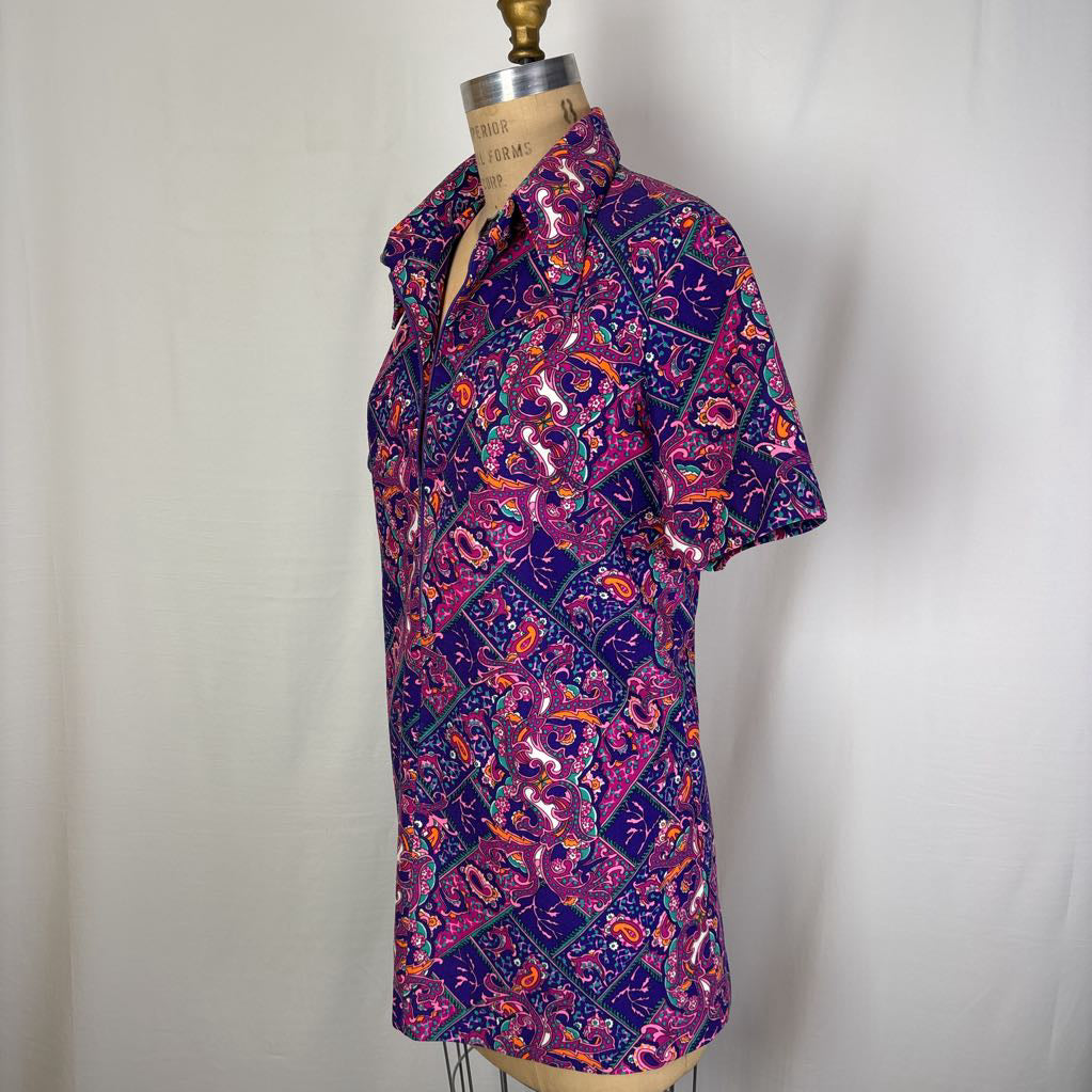 A short-sleeve button-up shirt with a vibrant purple, pink, and orange batik-style pattern, displayed on a dress form mannequin against a white fabric backdrop.