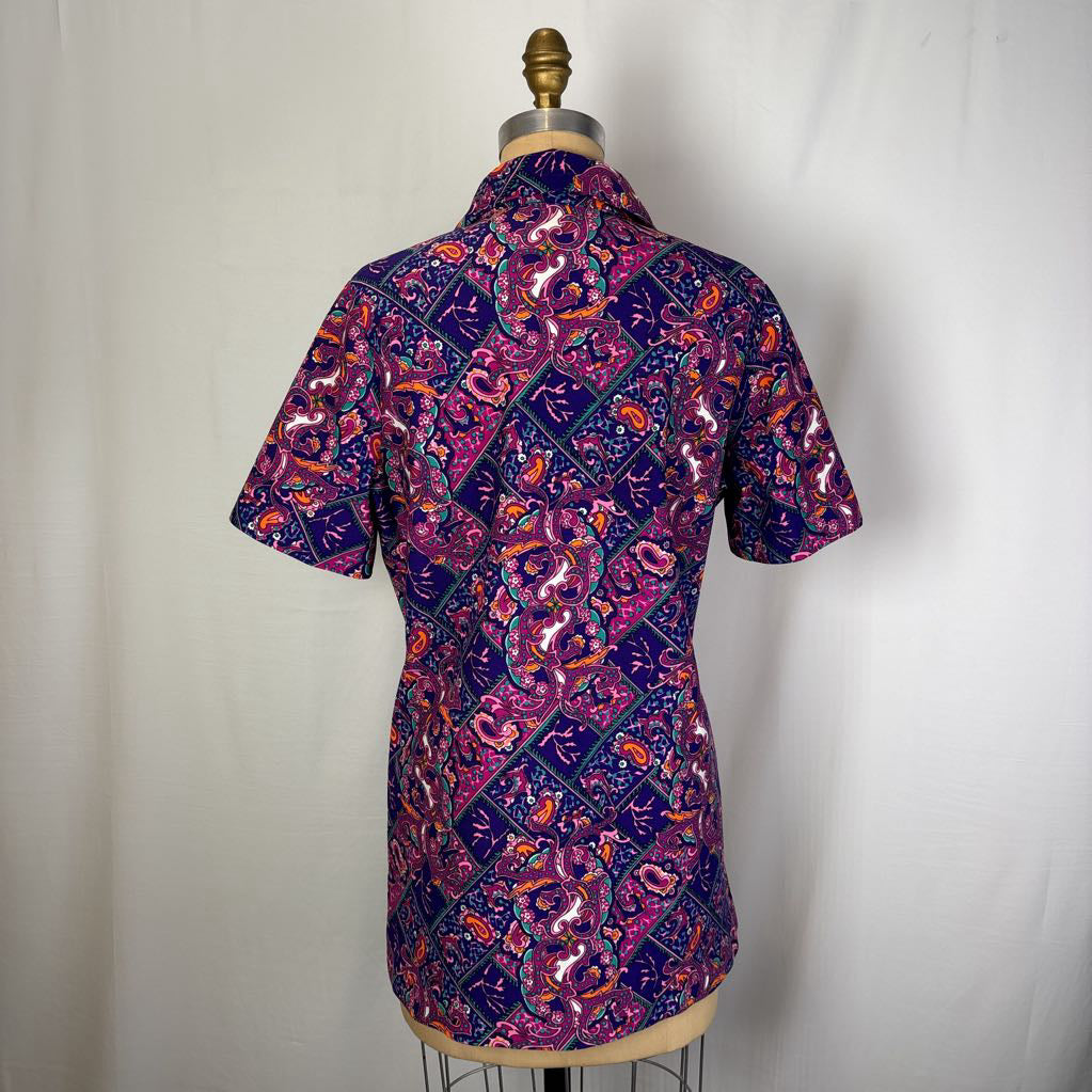 A short-sleeve button-up shirt with a vibrant batik pattern in purple, pink, orange, and blue, displayed on a dress form against a white fabric backdrop.